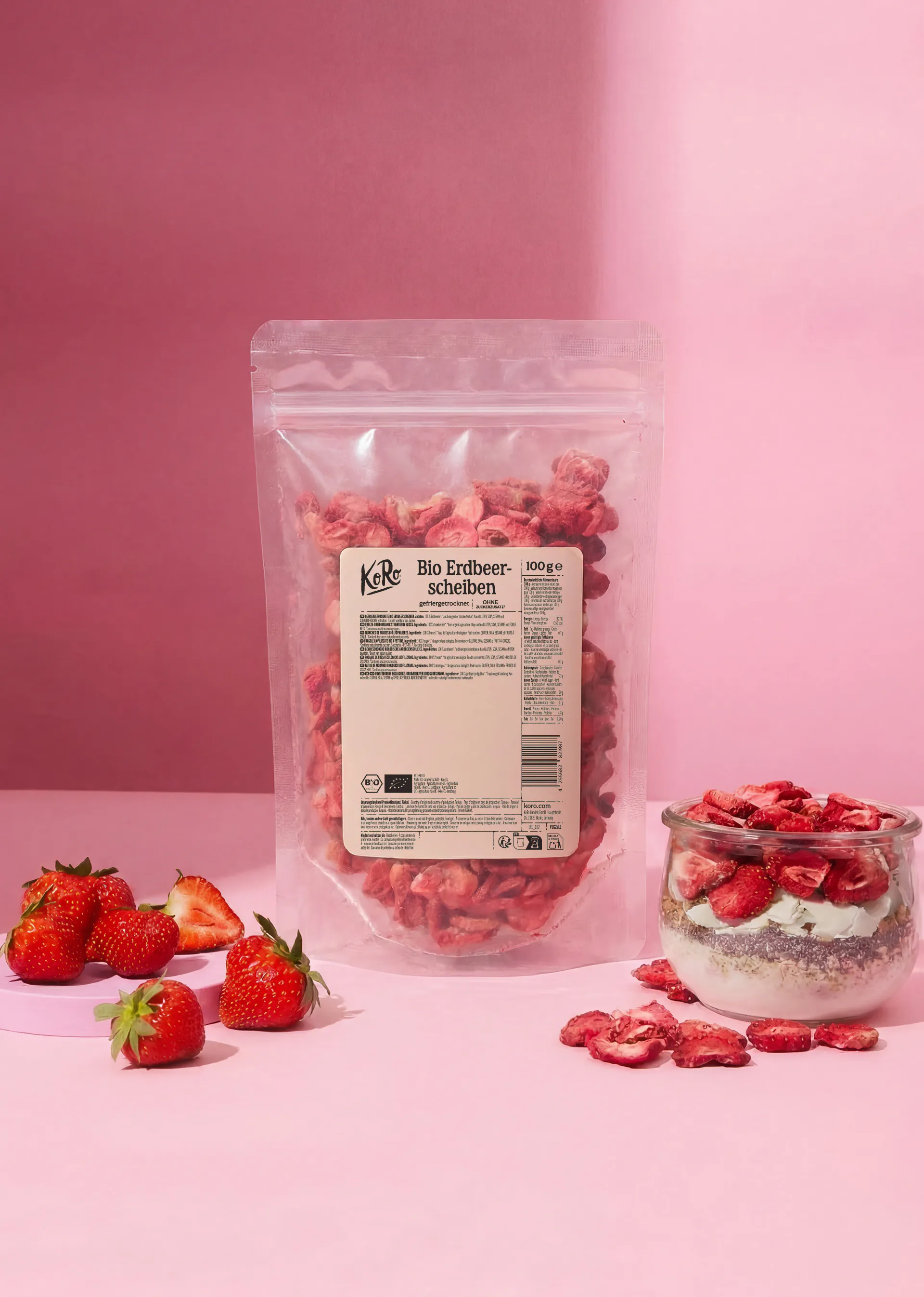 a bag of koro organic freeze-dried strawberry slices stands next to a glass jar of yogurt topped with the strawberries and a small pile of fresh strawberries on a pink background.