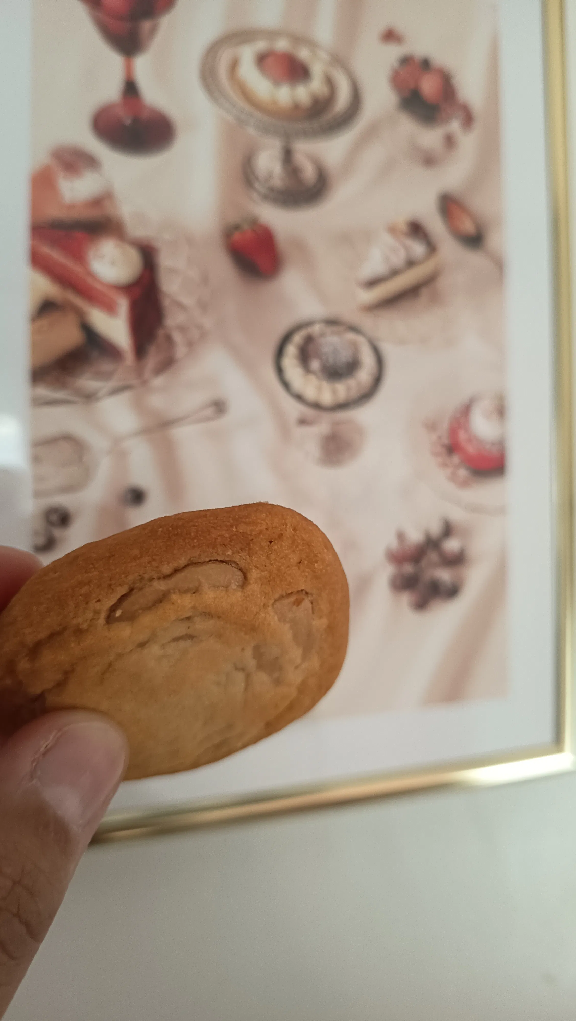 una mano tiene un biscotto rotondo in primo piano davanti a un quadro raffigurante vari dolci e dessert.