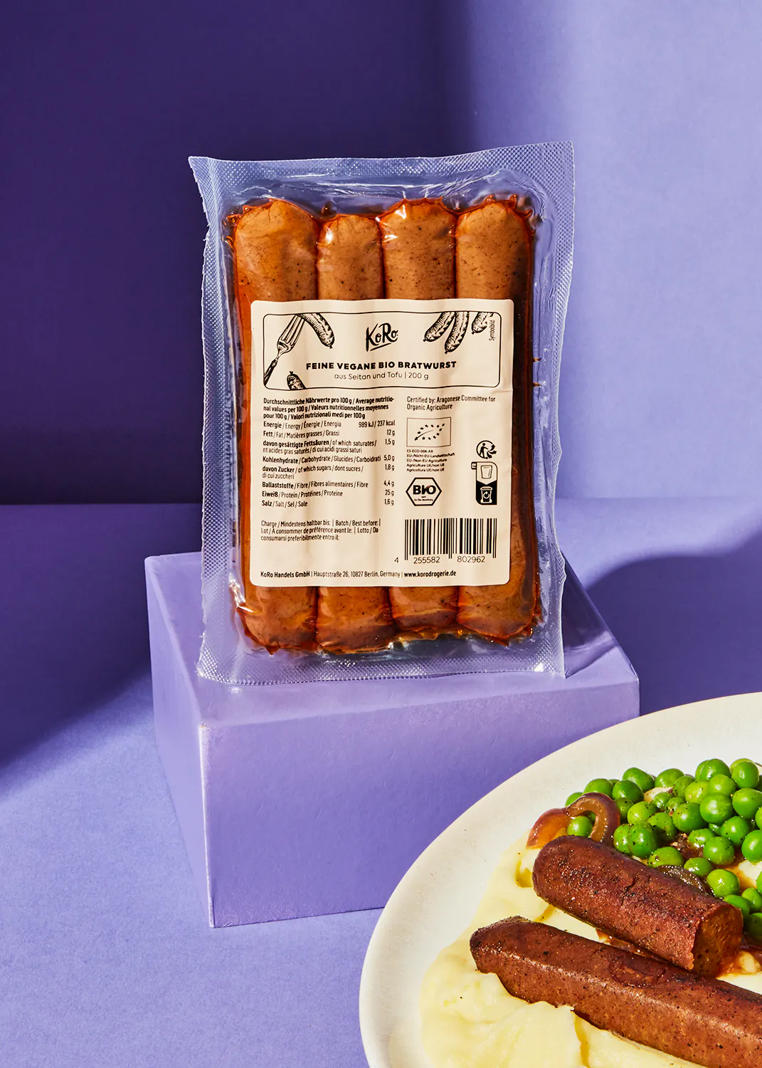 un sac de saucisses végétales est assis sur une table violette à côté d' une assiette de pommes de terre