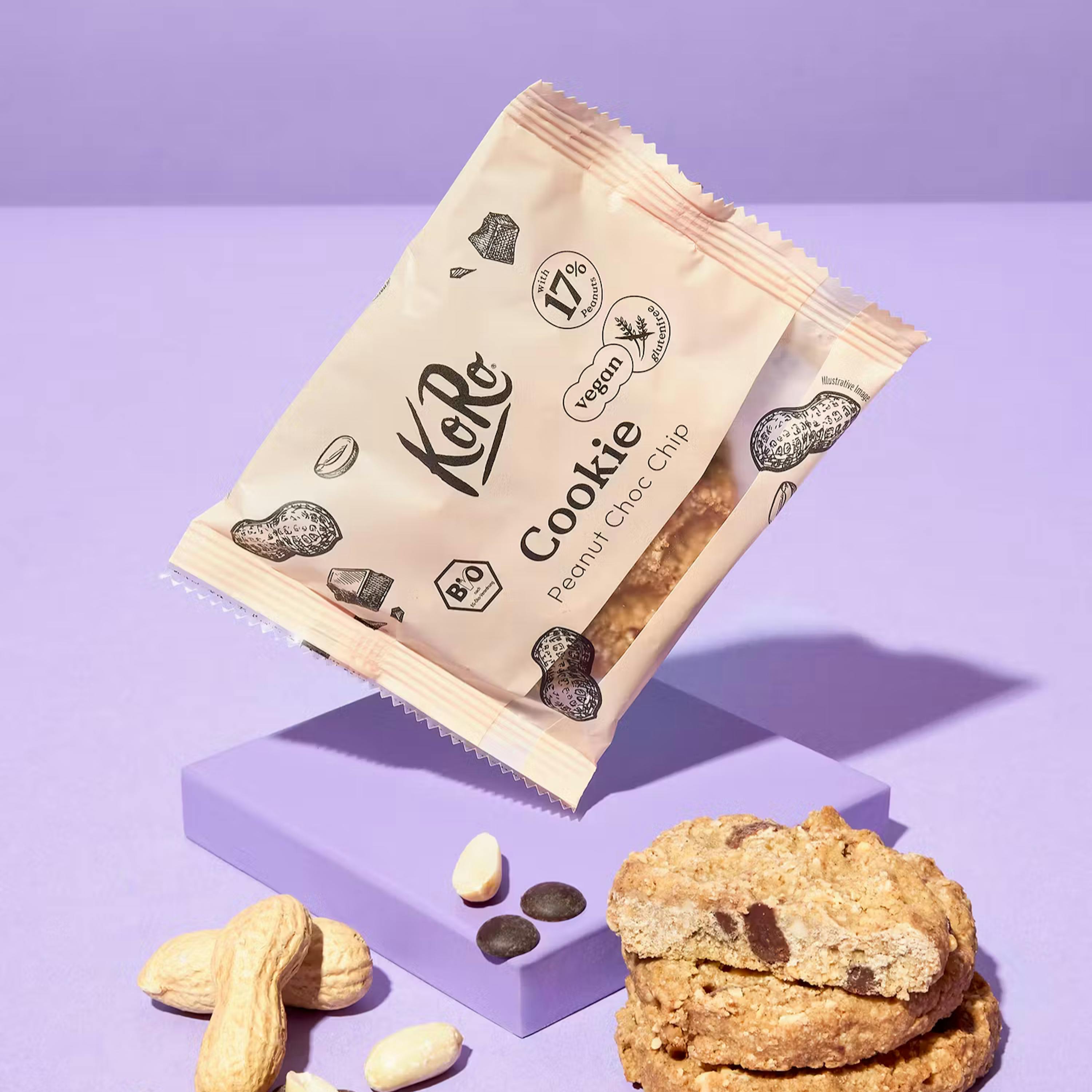 un pacchetto di biscotti koro al burro d'arachidi e gocce di cioccolato è posizionato sopra un blocco viola, con arachidi e gocce di cioccolato sparse e due biscotti impilati, uno dei quali spezzato a metà, su uno sfondo viola.