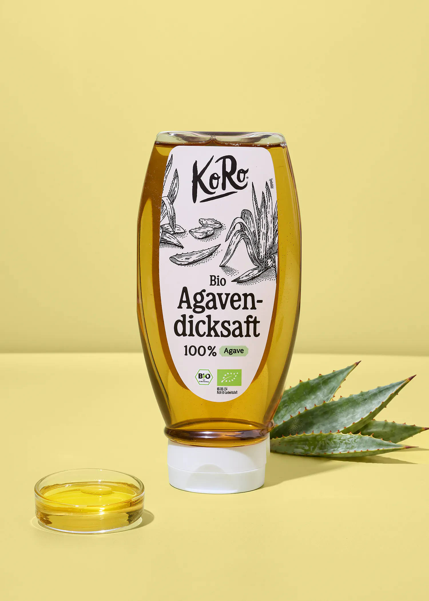 bouteille de sirop d'agave bio koro posée sur un fond jaune avec une coupelle de sirop et des feuilles d'agave à côté.