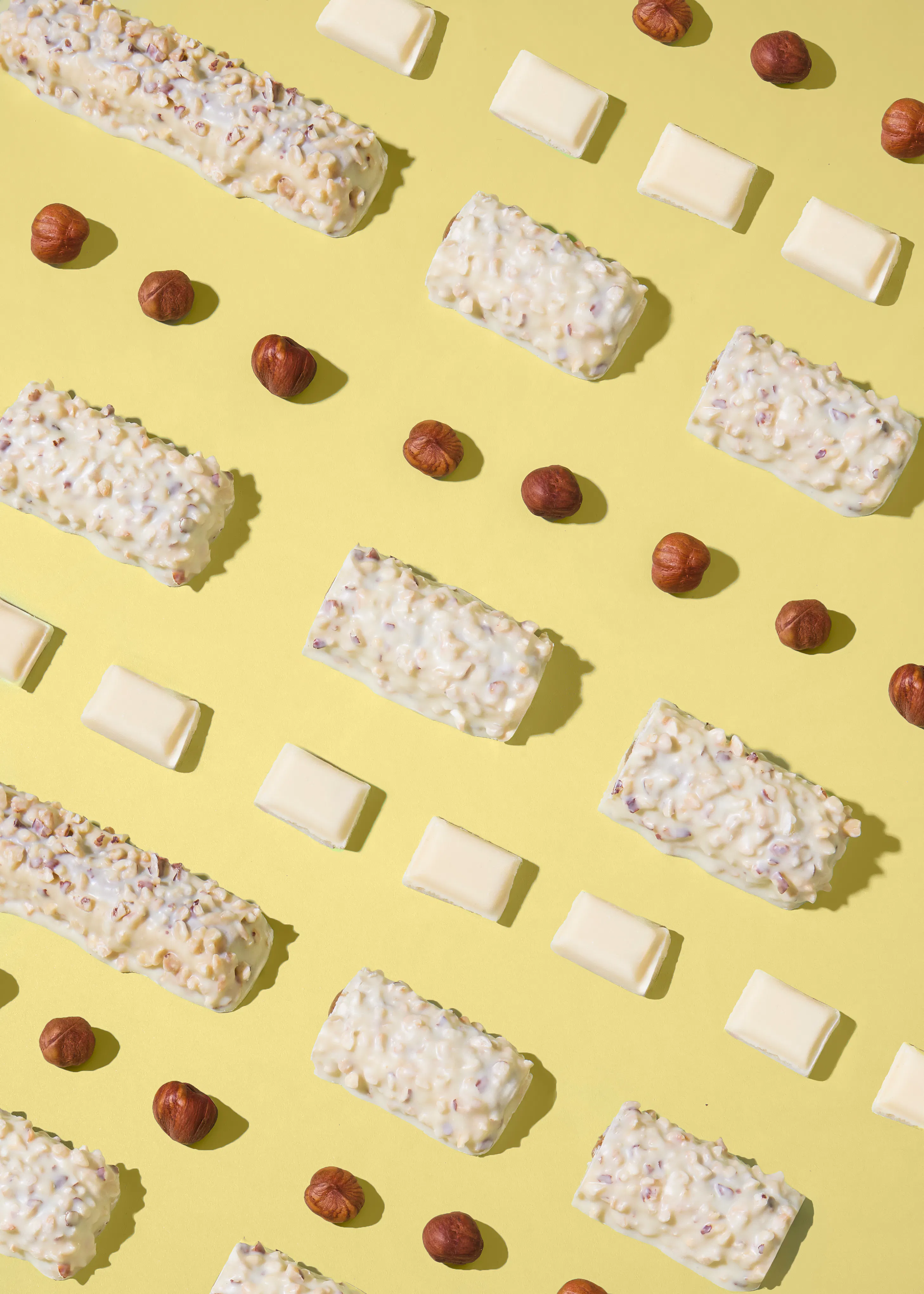 a flat lay pattern of white chocolate bars with nut pieces, whole hazelnuts, and small white chocolate squares on a yellow background.