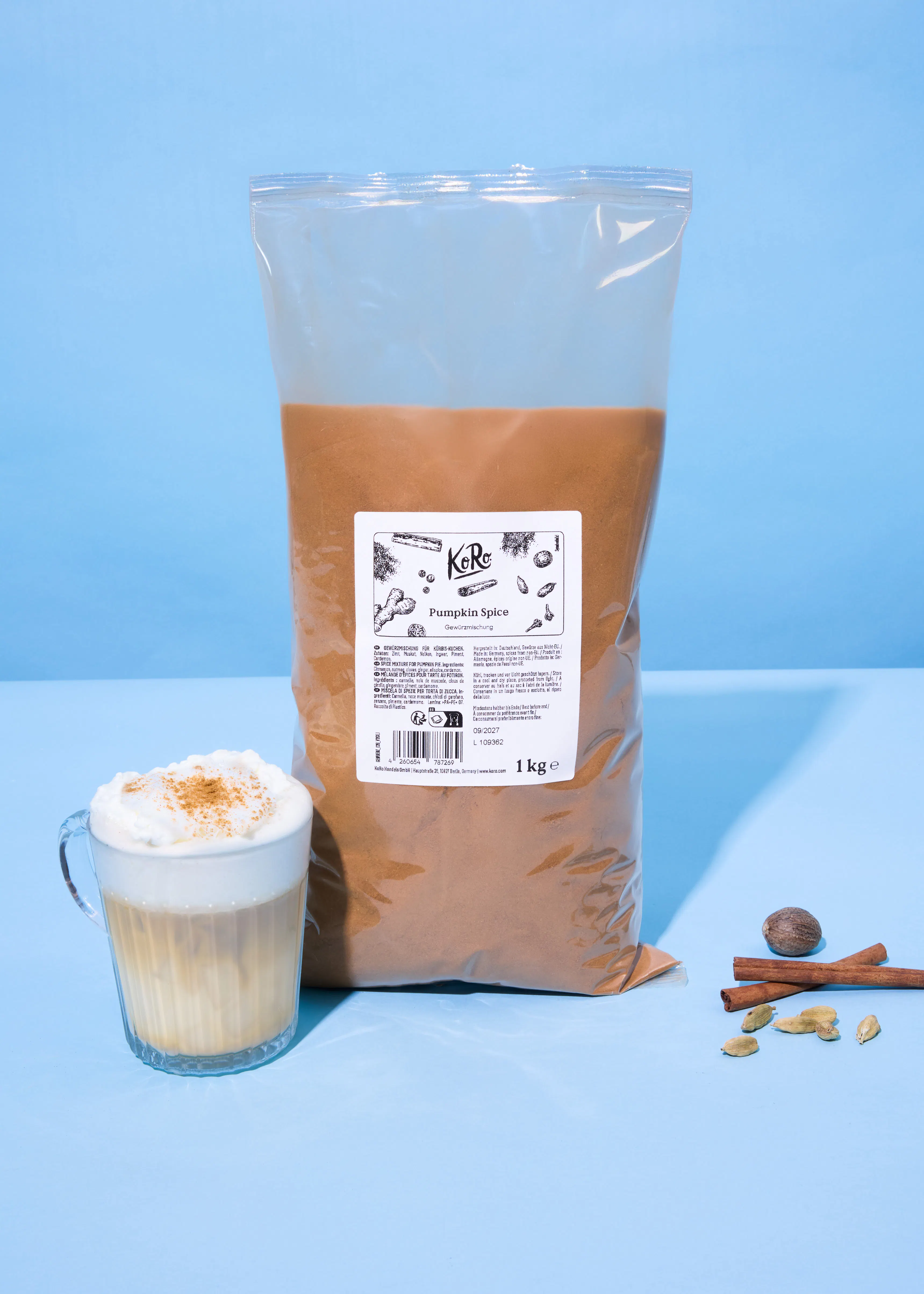 un grand sac d'épices pour citrouille, un latte aux épices pour citrouille et des épices entières (muscade, cannelle, cardamome) sont présentés sur un fond bleu.