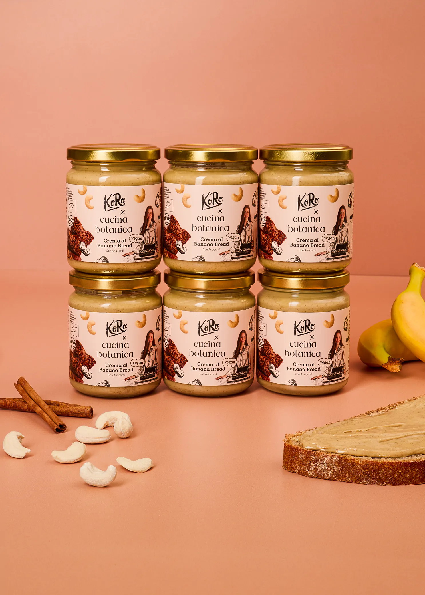 sechs gläser koro x cucina botanica banana bread creme auf einem pfirsichfarbenen hintergrund, umgeben von bananen, cashews, zimtstangen und einer bestrichenen brotscheibe.