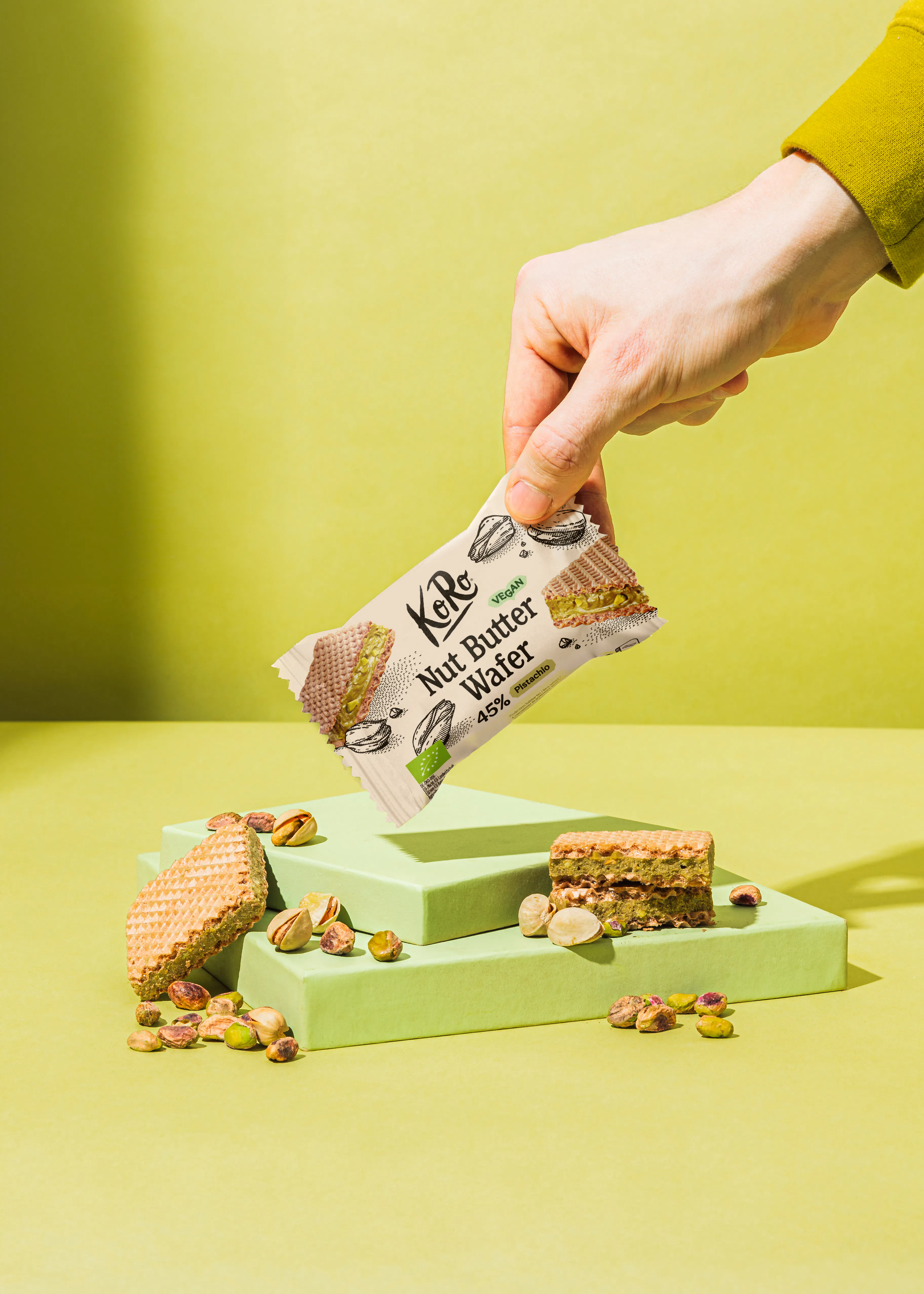 eine hand hält eine koro nut butter wafer waffel über hellgrünen podesten, die mit pistazien und waffelstücken dekoriert sind.