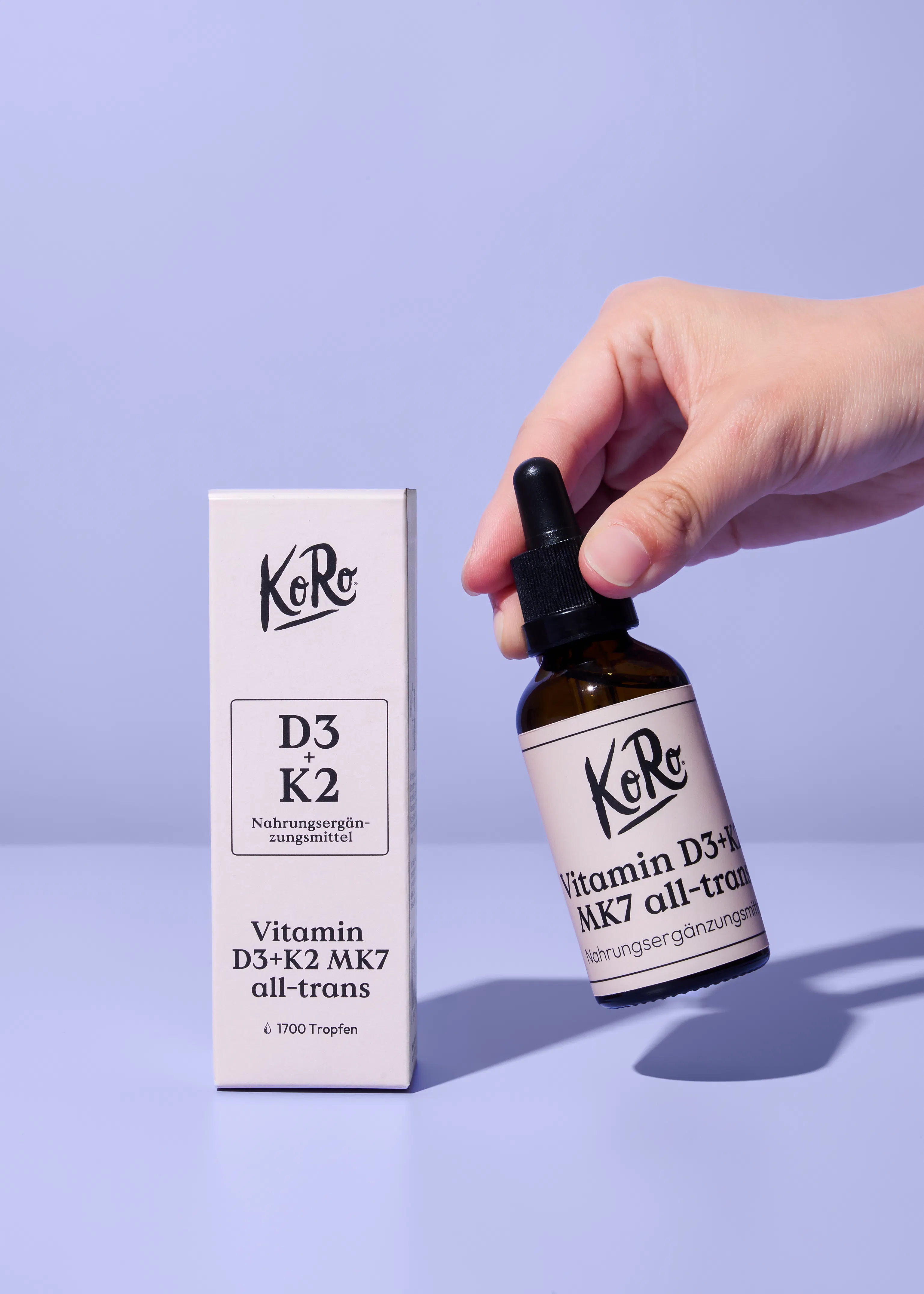 a hand holds a koro vitamin d3+k2 mk7 all-trans dropper bottle next to its matching box on a light purple background.