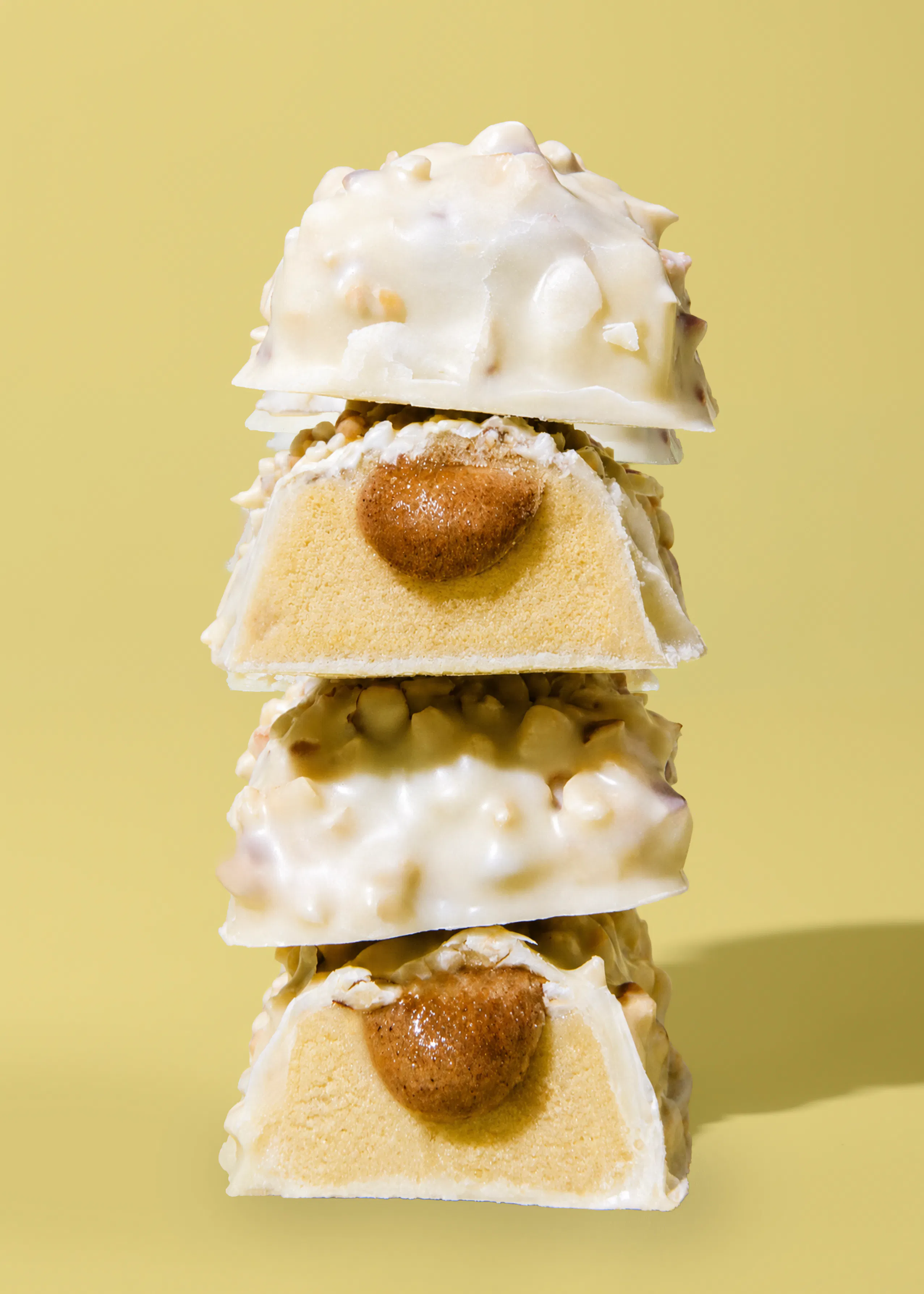 une pile de quatre friandises au chocolat blanc parsemées d'éclats, dont deux sont coupées en deux pour révéler un centre de caramel ou de pâte à tartiner sur une base claire, le tout sur un fond jaune uni.