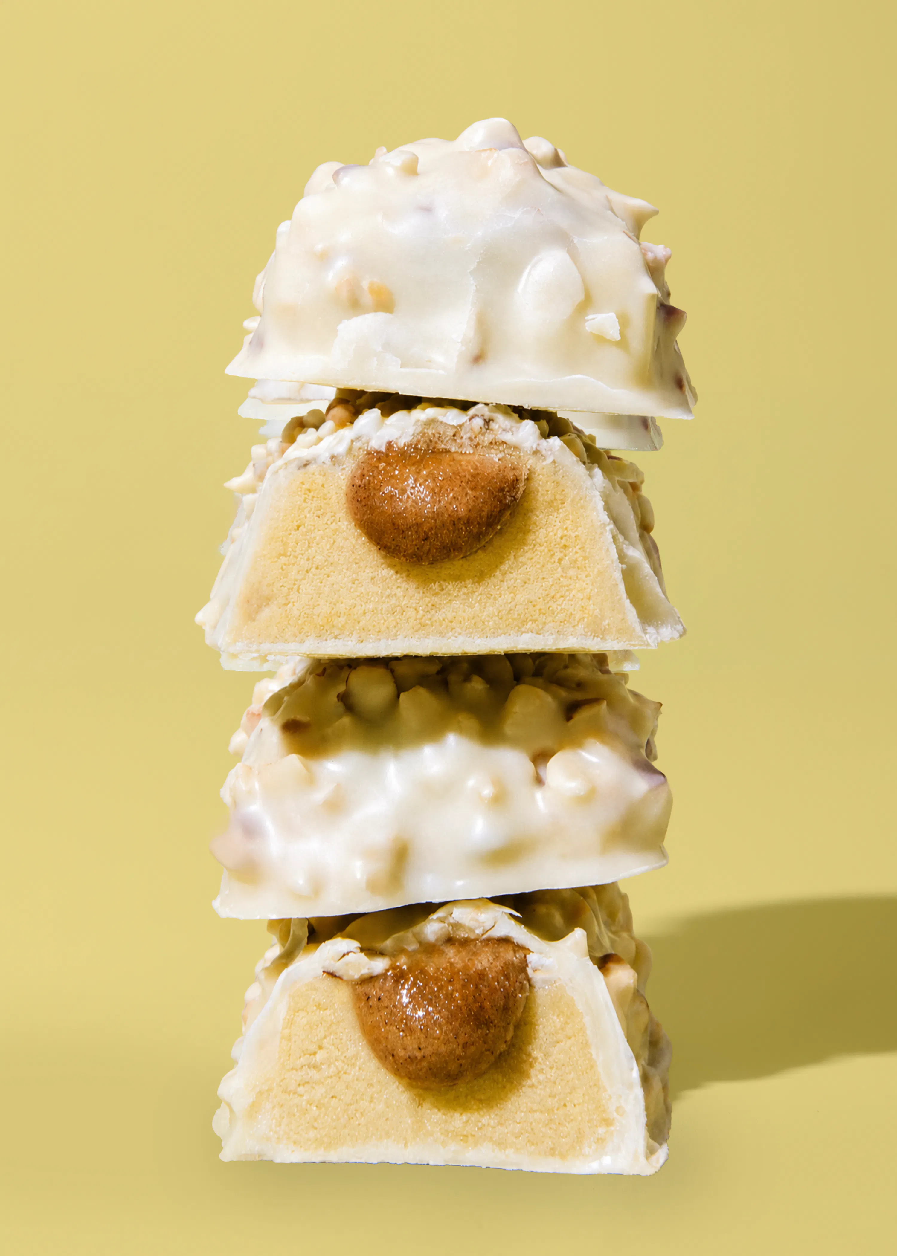 une pile de quatre friandises au chocolat blanc parsemées d'éclats, dont deux sont coupées en deux pour révéler un centre de caramel ou de pâte à tartiner sur une base claire, le tout sur un fond jaune uni.