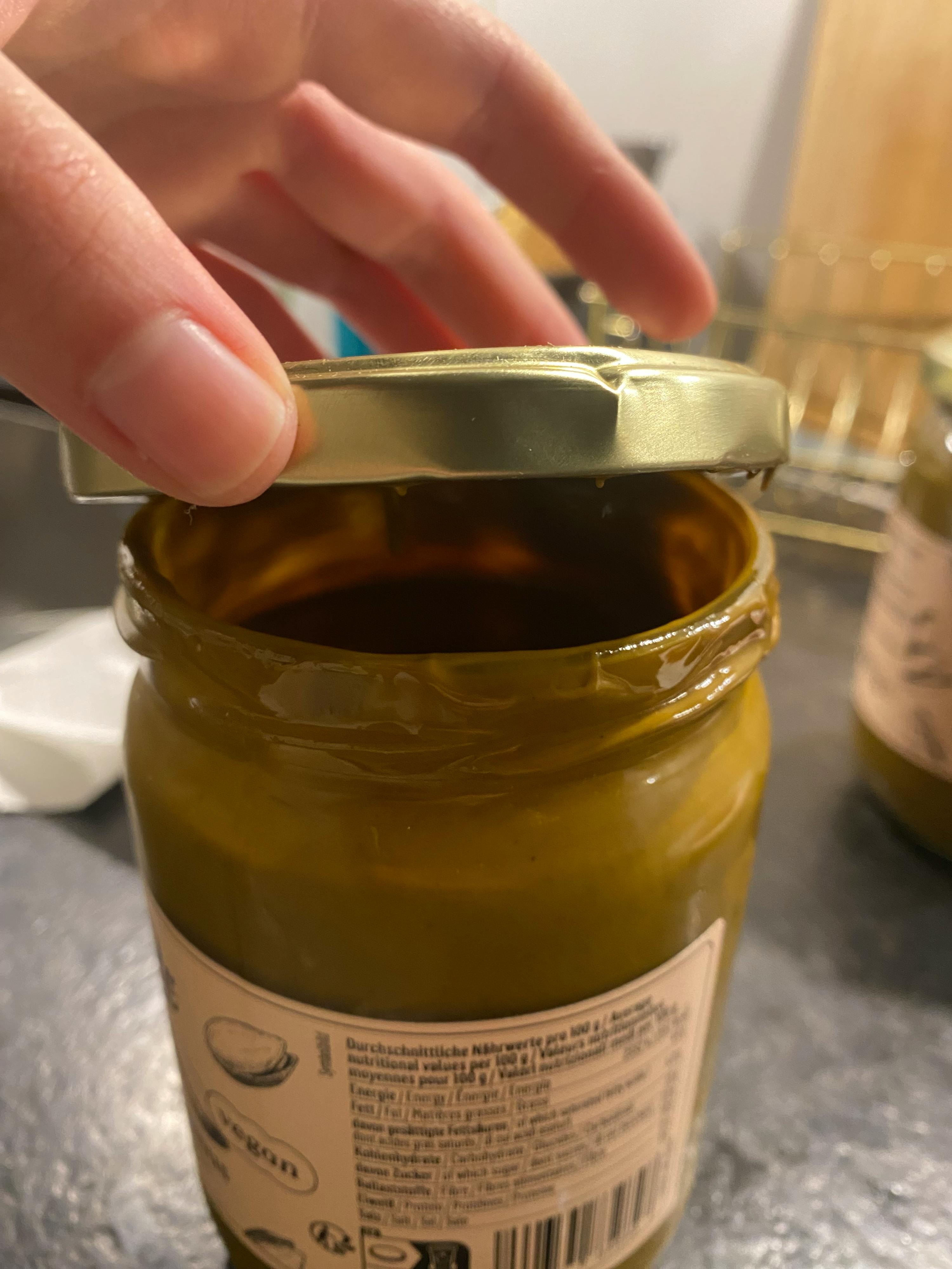 a hand holds an open jar of greenish-brown spread with a label showing nutritional values and the word 'vegan'.