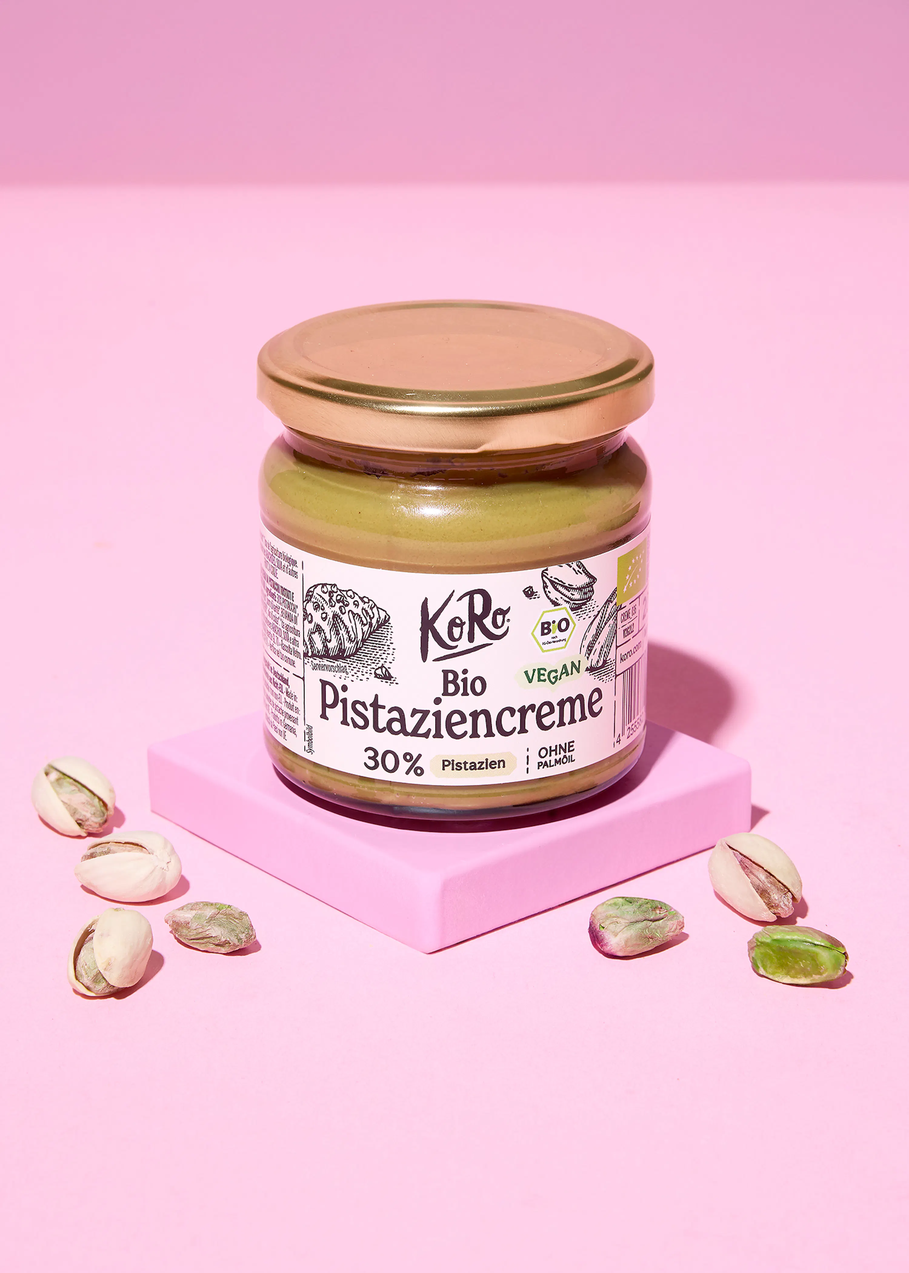 a koro bio vegan pistachio cream jar with 30% pistachios and no palm oil, surrounded by pistachios, sits on a pink stand against a pink background.