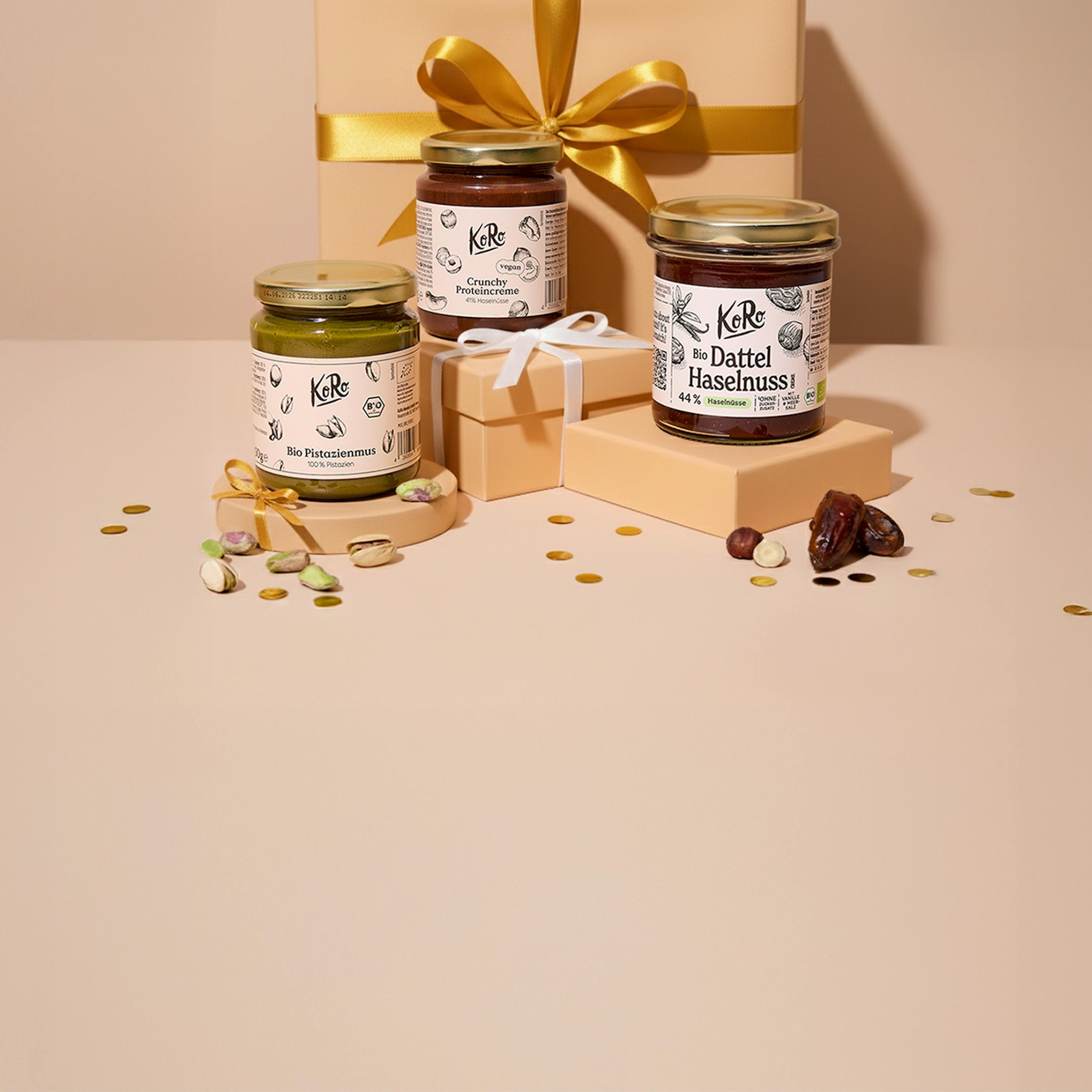 three jars of koro nut butter, including pistachio, crunchy protein, and date hazelnut, are arranged with gift boxes, loose nuts, and gold confetti on a light background.