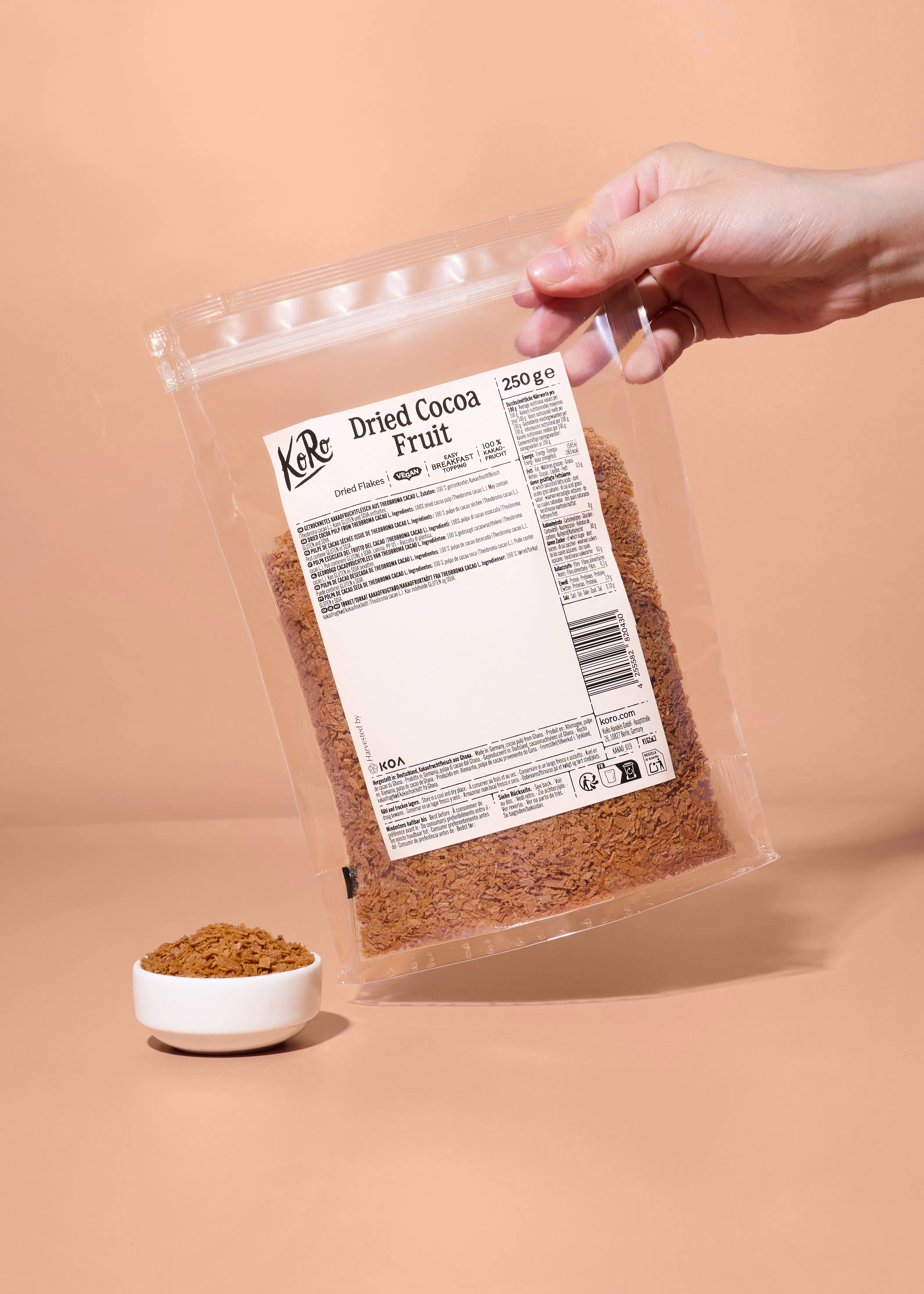 eine hand hält eine durchsichtige verpackung mit getrocknetem kakaofruchtfleisch von koro über einer kleinen weißen schale mit dem inhalt.