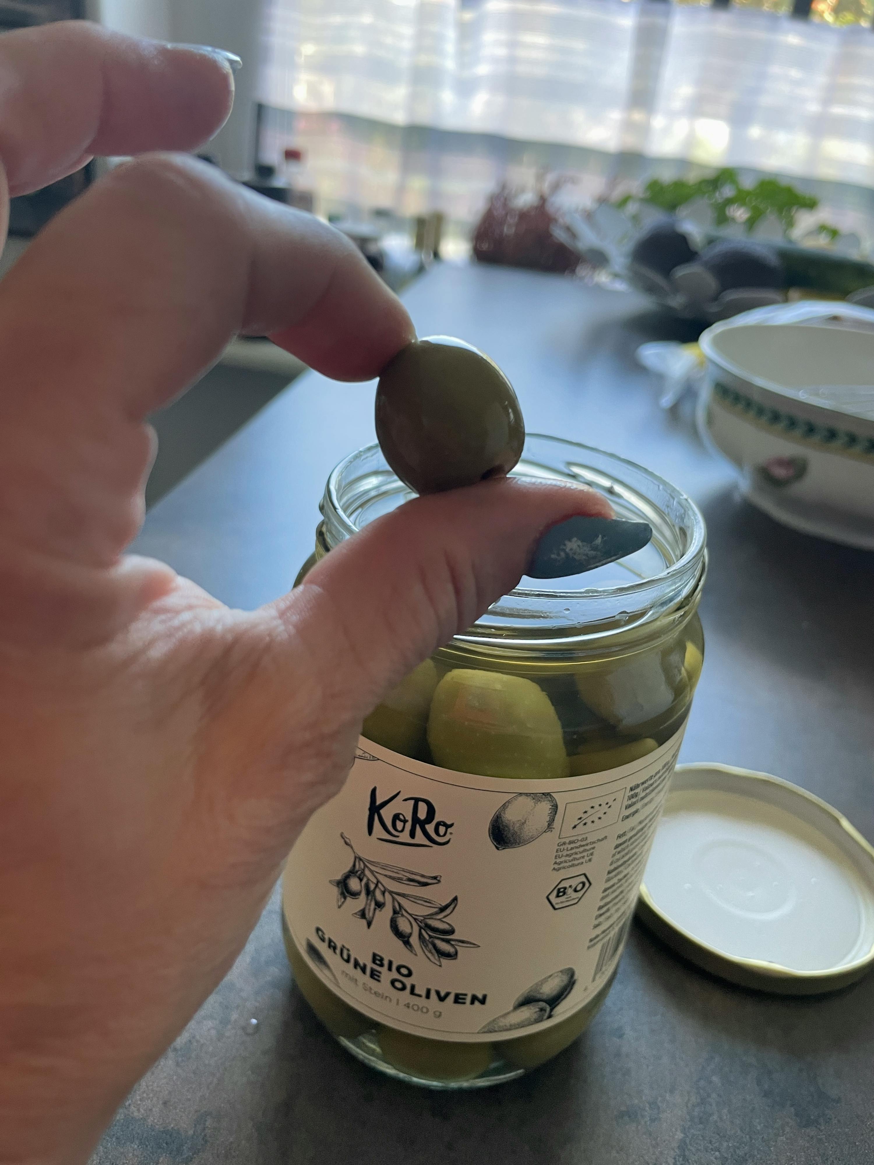 a person holds a green olive in front of a jar of organic olives