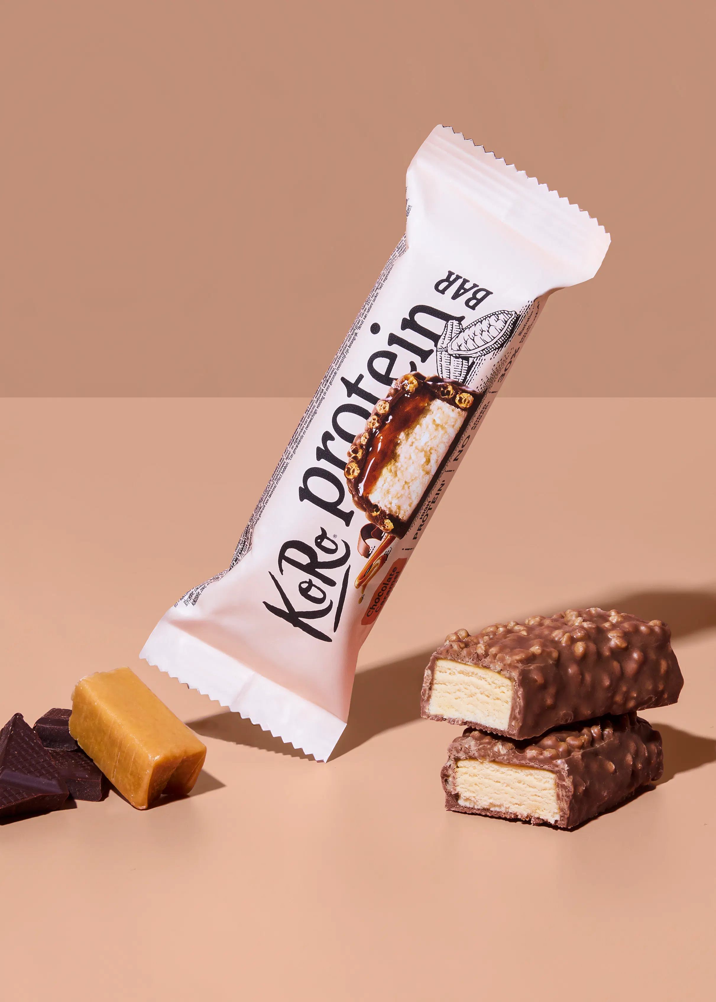 a koro protein bar in its wrapper, two chocolate-covered protein bars with creamy filling, a piece of caramel, and chocolate chunks on a beige background.
