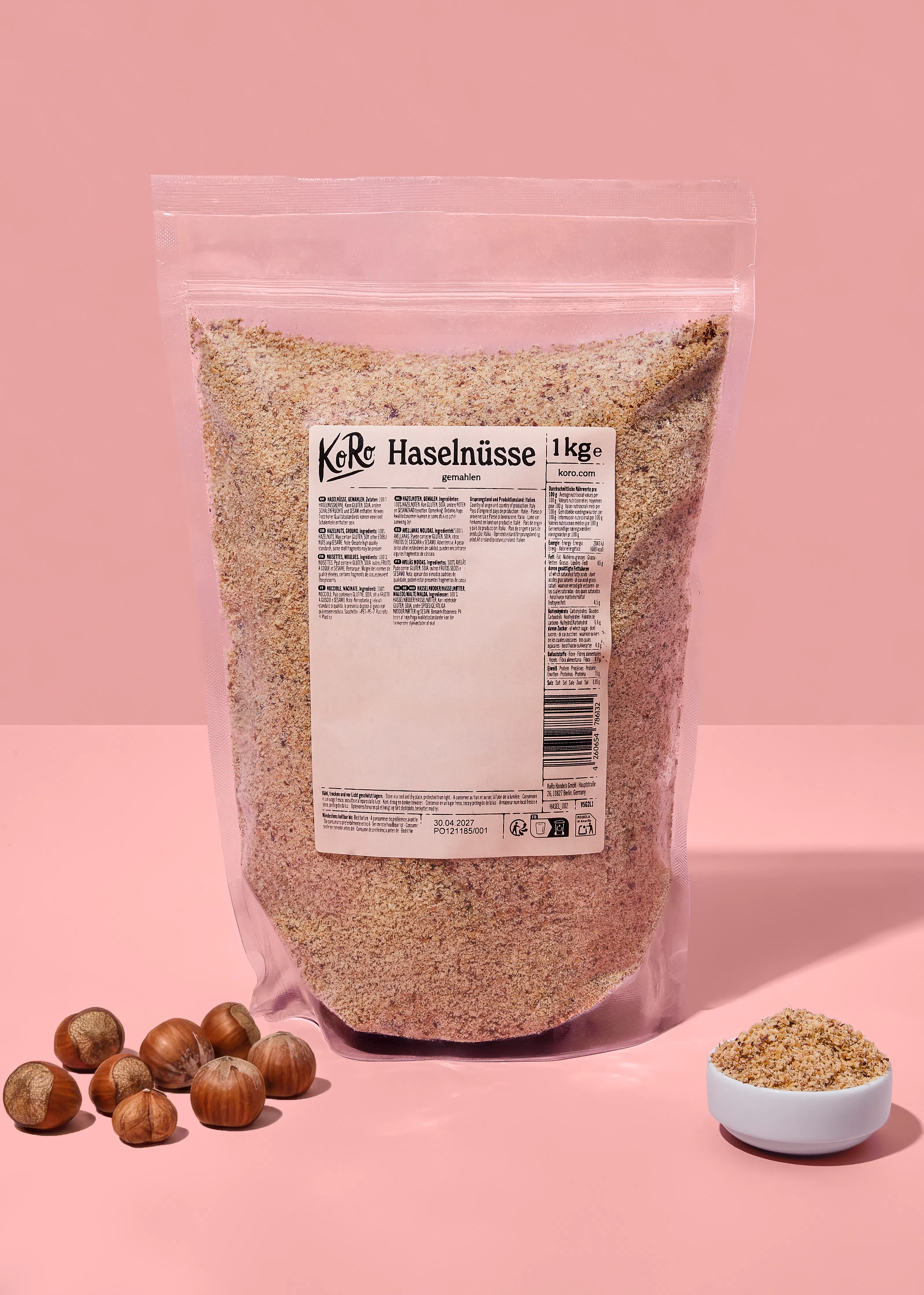 eine große, durchsichtige tüte mit 1 kg gemahlenen haselnüssen der marke koro steht auf einem rosafarbenen hintergrund, daneben liegen ganze haselnüsse in der schale und eine kleine schale mit gemahlenen haselnüssen.