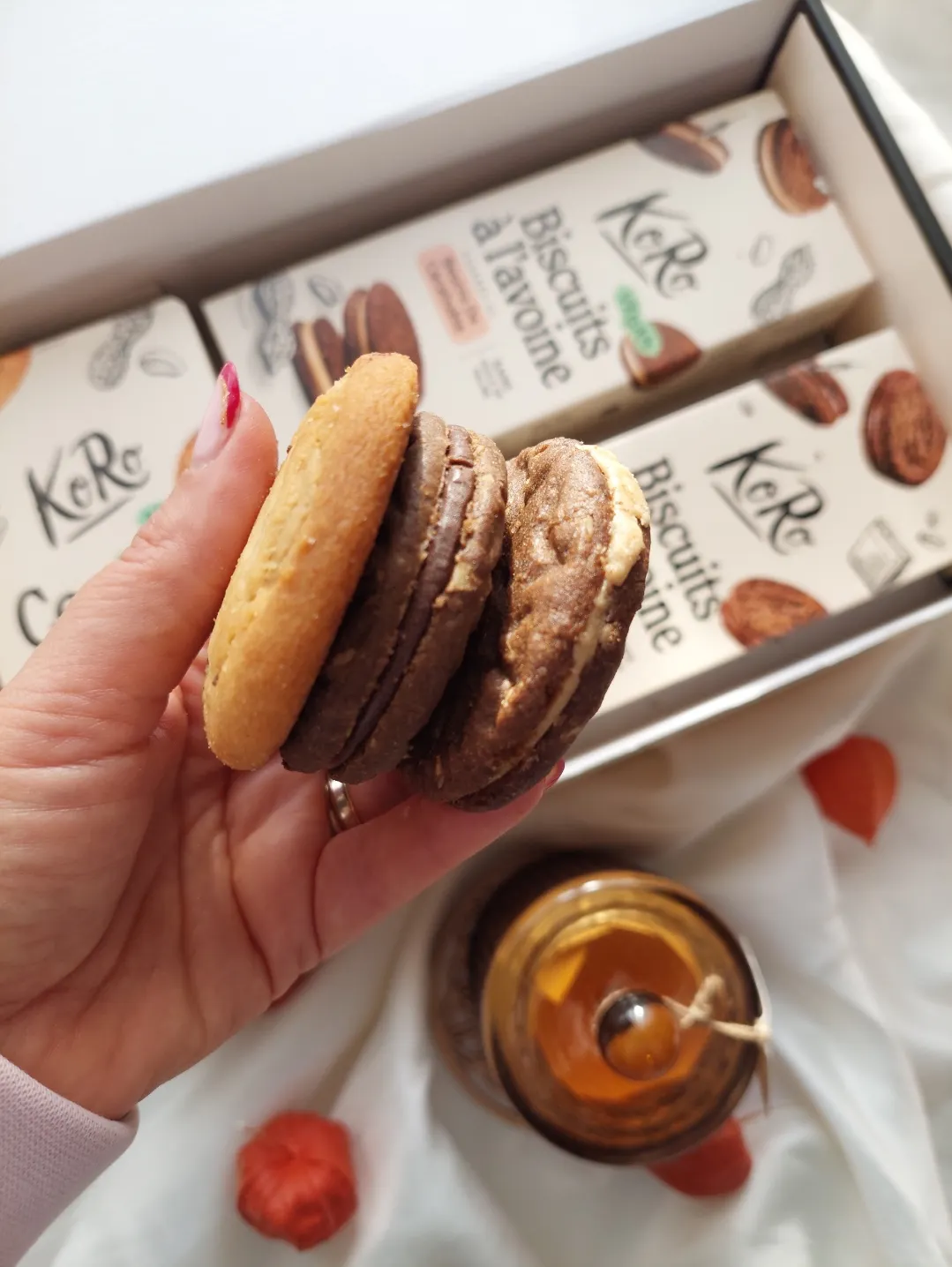 una mano tiene tre biscotti farciti di diversi gusti davanti a una scatola di biscotti koro e un vasetto di miele.