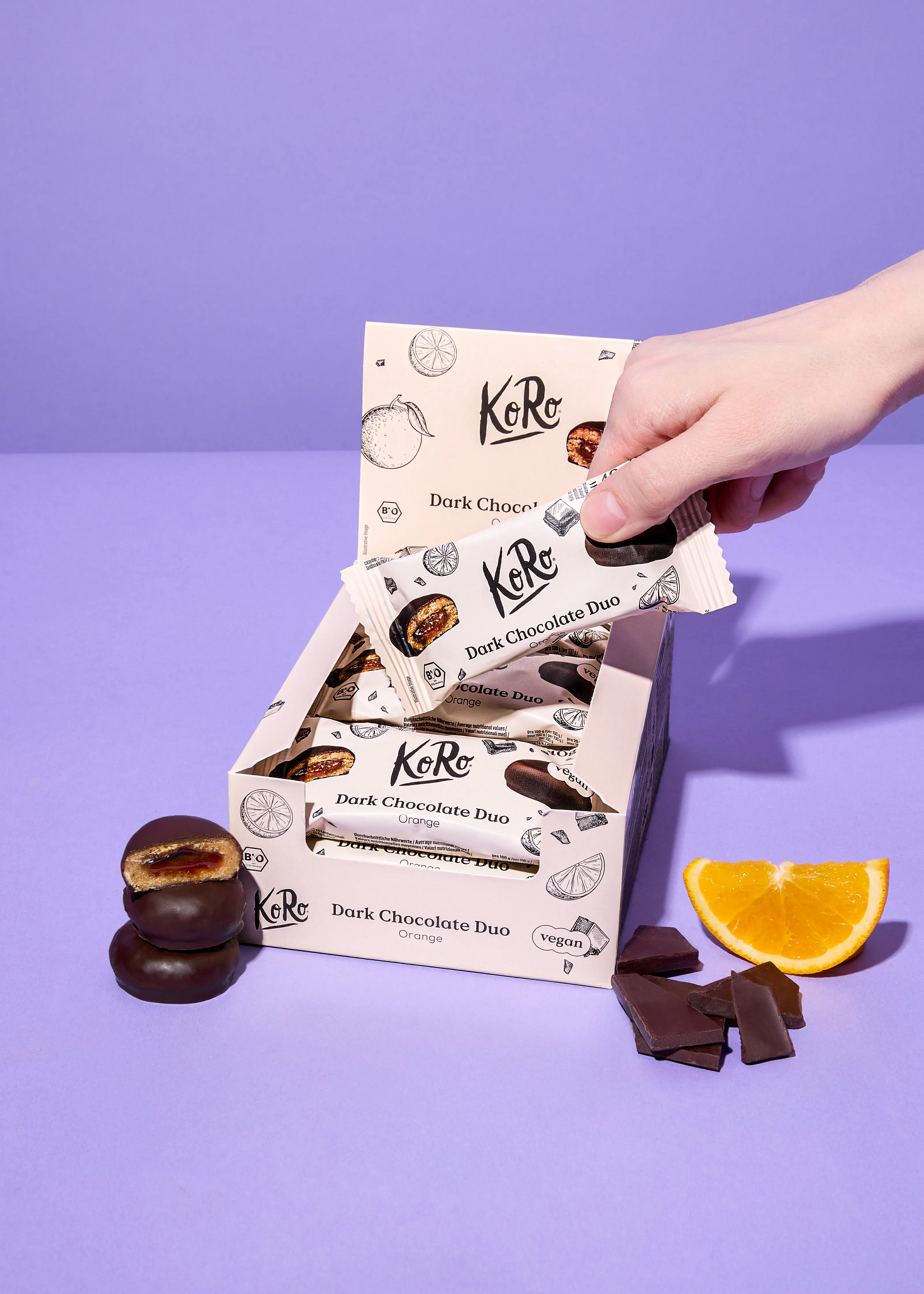 a hand reaches into a box of koro dark chocolate duo orange vegan snacks, with loose snacks, chocolate pieces, and an orange slice on a purple background.