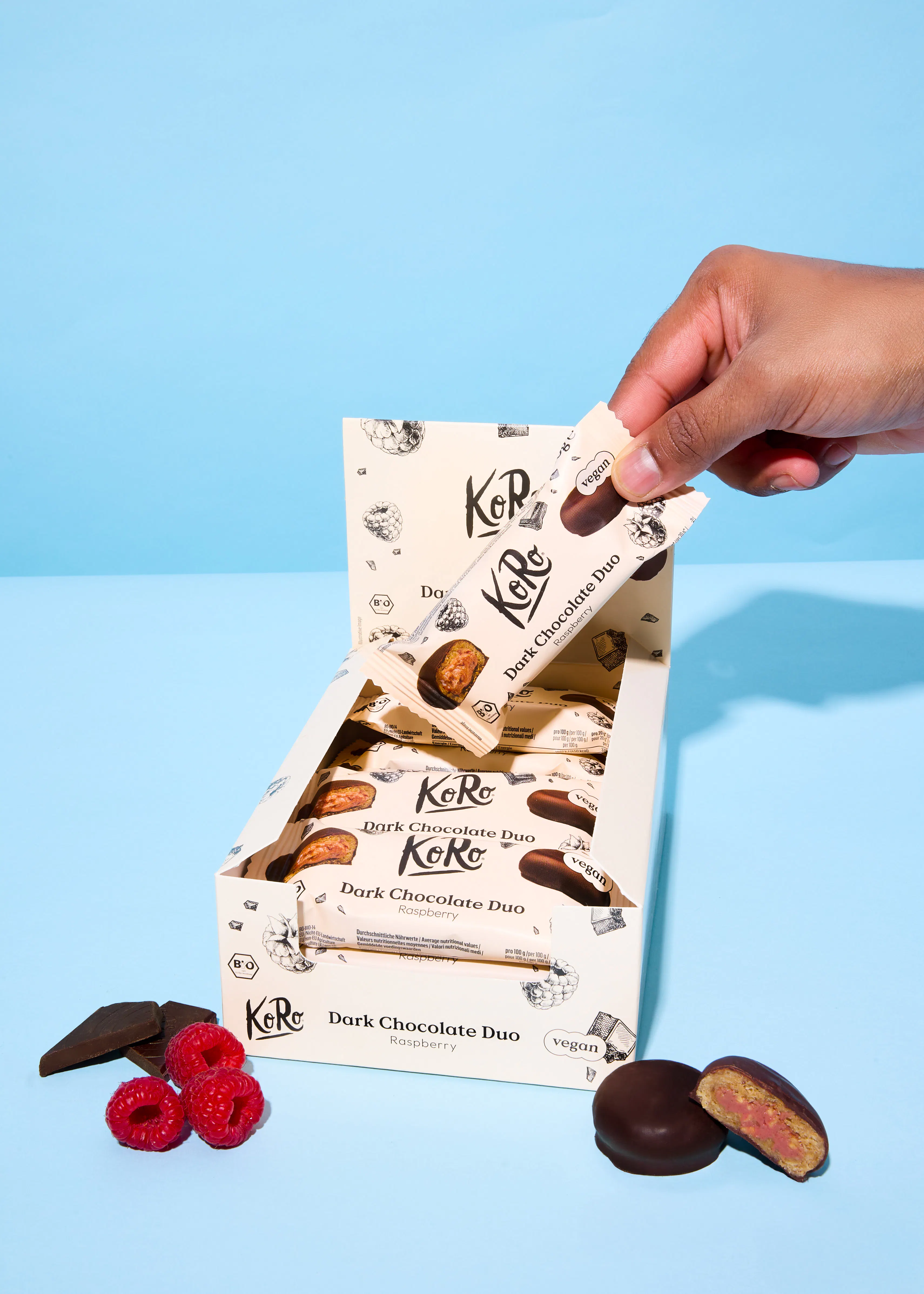 a hand reaches into an open box of koro dark chocolate duo raspberry bars, with loose raspberries, a chocolate piece, and a cut bar revealing a pink filling on a blue surface.