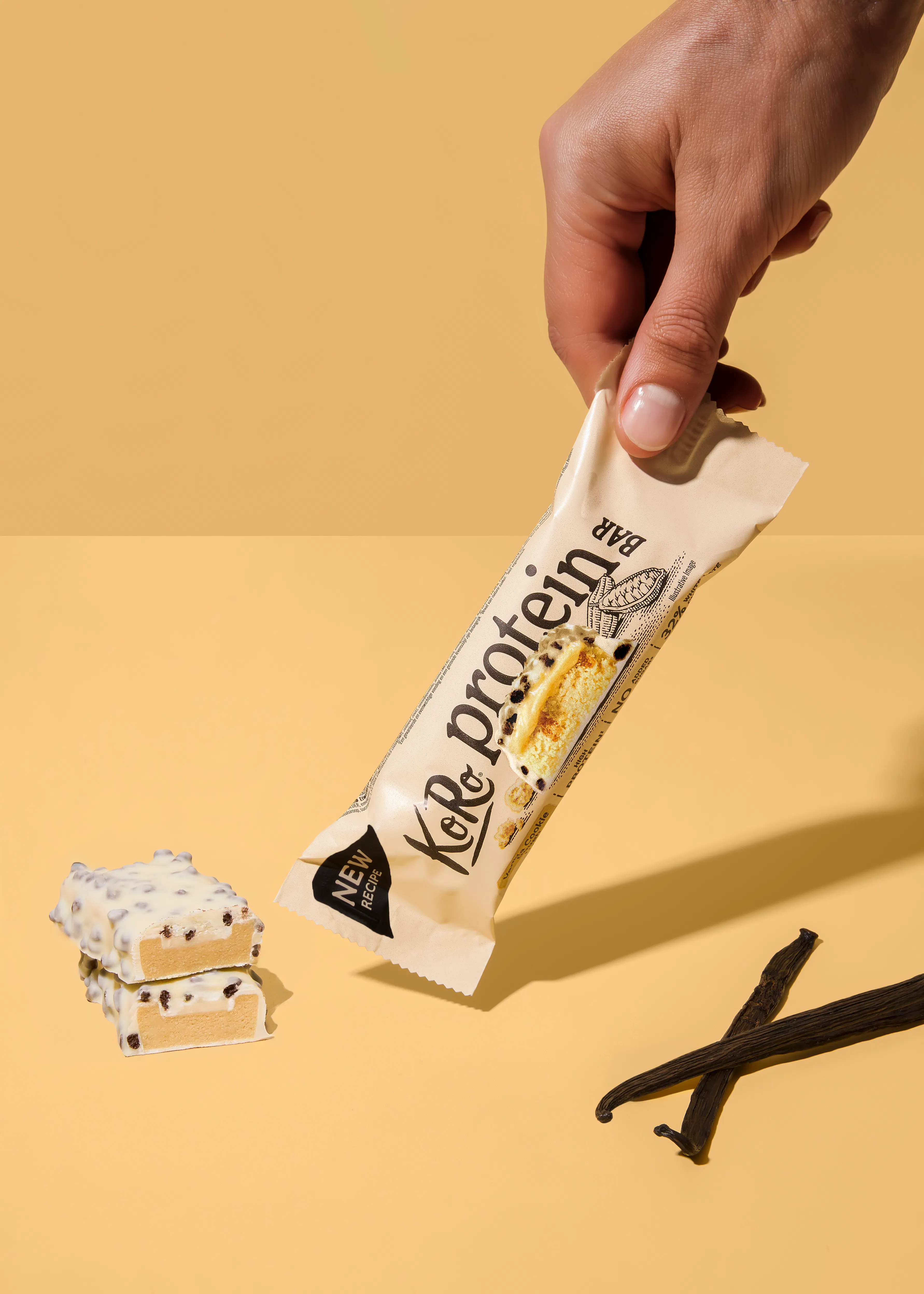 eine hand hält einen koro proteinriegel mit vanille-cookie-geschmack, daneben liegen zwei angeschnittene riegel und vanilleschoten auf gelbem hintergrund.