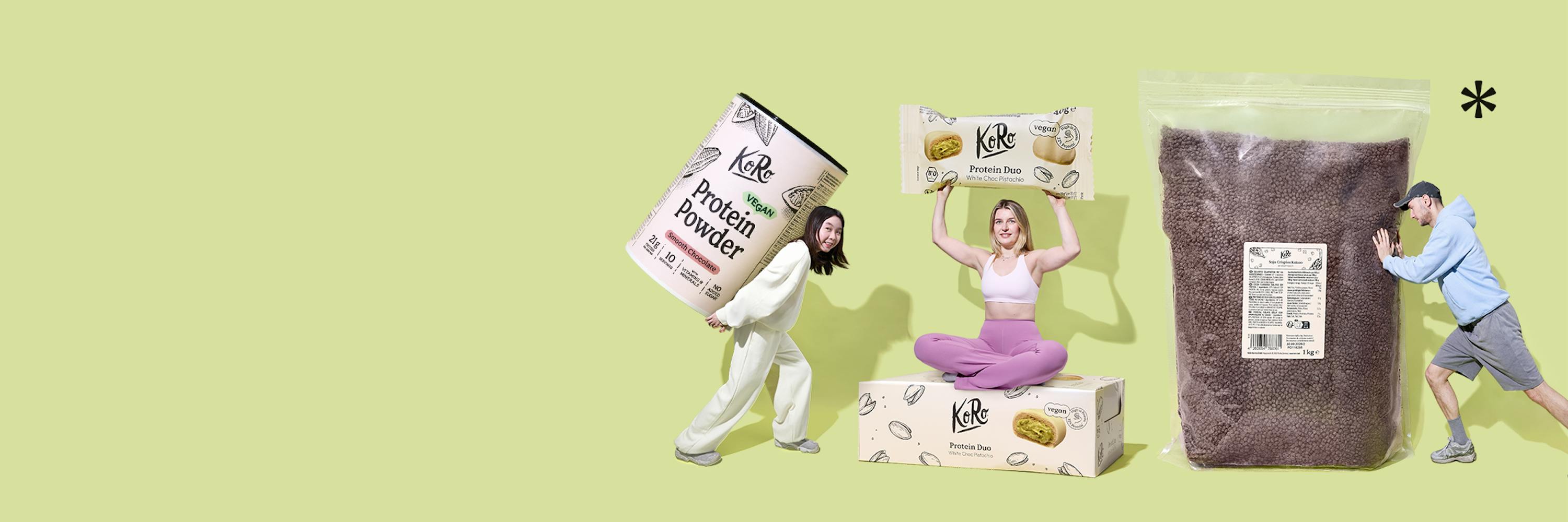 tre persone interagiscono con prodotti koro di grandi dimensioni: una donna trasporta un barattolo di proteine in polvere, un'altra tiene in alto una barretta proteica e un uomo spinge un grande sacco di prodotto sfuso.