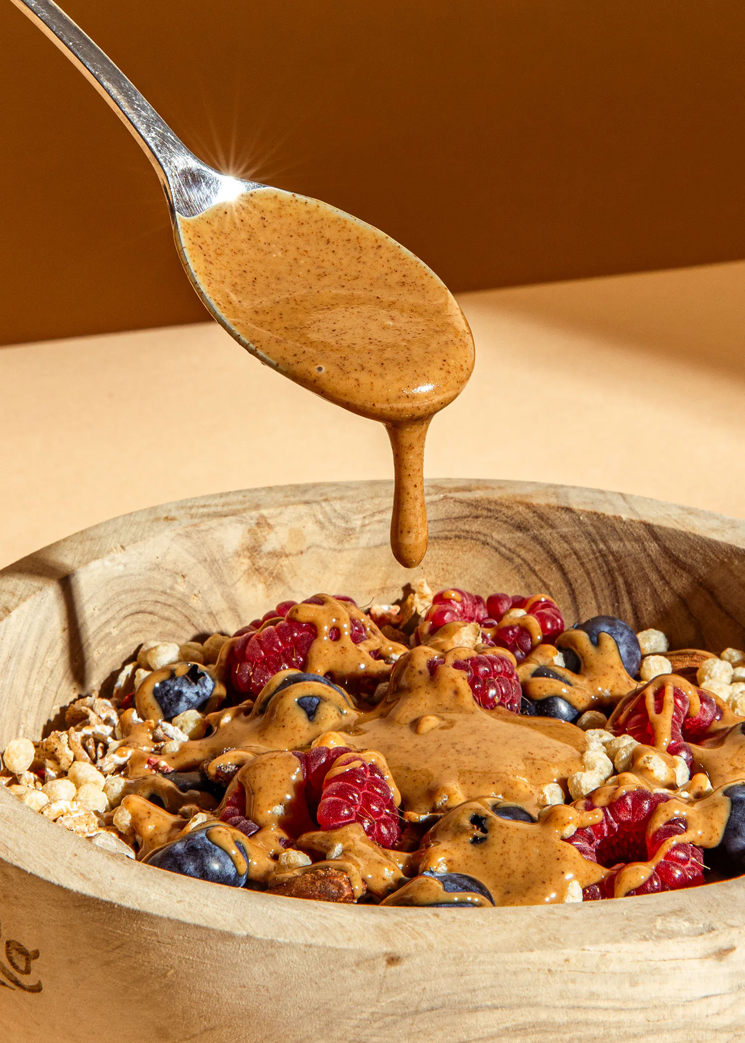 une cuillère verse du beurre d' arachide sur un bol en bois rempli de fruits et de granola