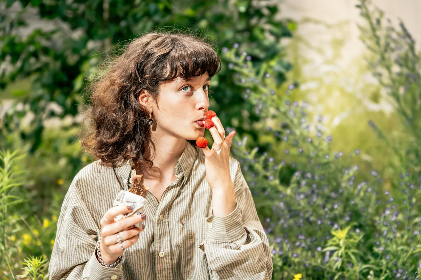 eine junge frau mit lockigem haar in einem gestreiften hemd hält eine schokoladentafel und isst erdbeeren vor einem unscharfen hintergrund aus grünpflanzen und blumen.