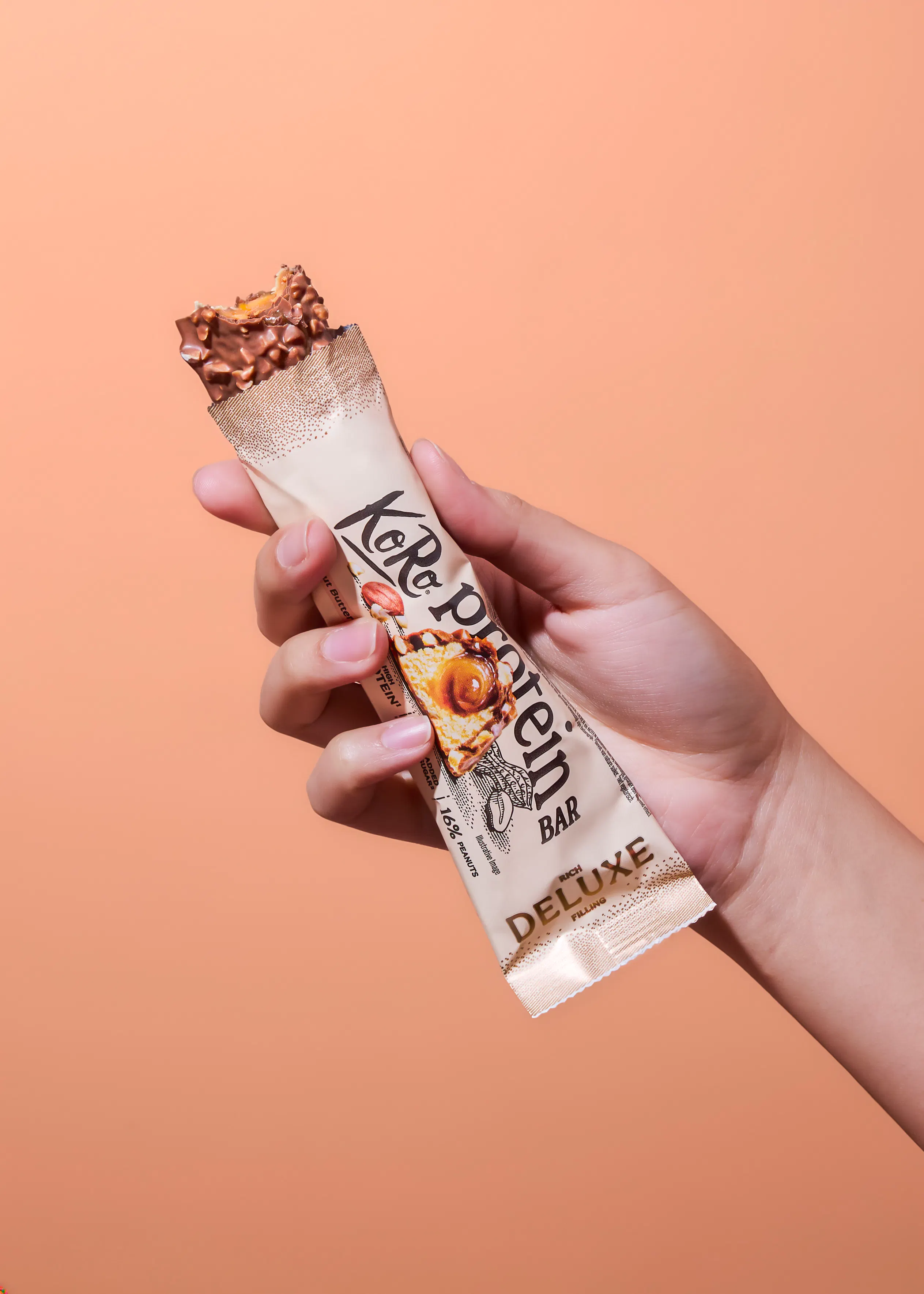 a hand holds a partially opened koro protein bar, revealing its chocolate and nut coating, against a plain background.