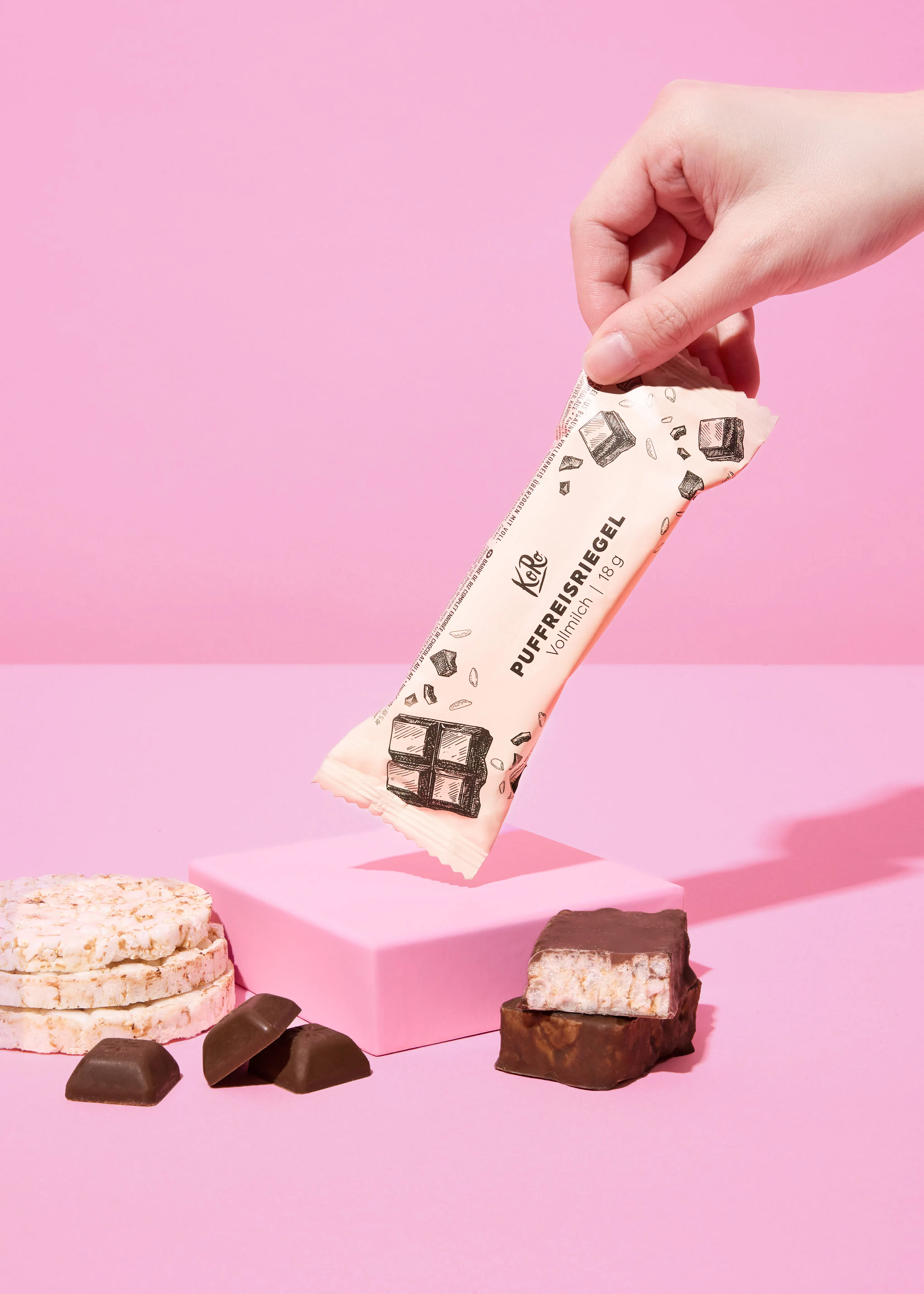 une main tient une barre de riz soufflé emballée au-dessus d'une surface rose, à côté de galettes de riz, de morceaux de chocolat et d'une barre de riz soufflé coupée.