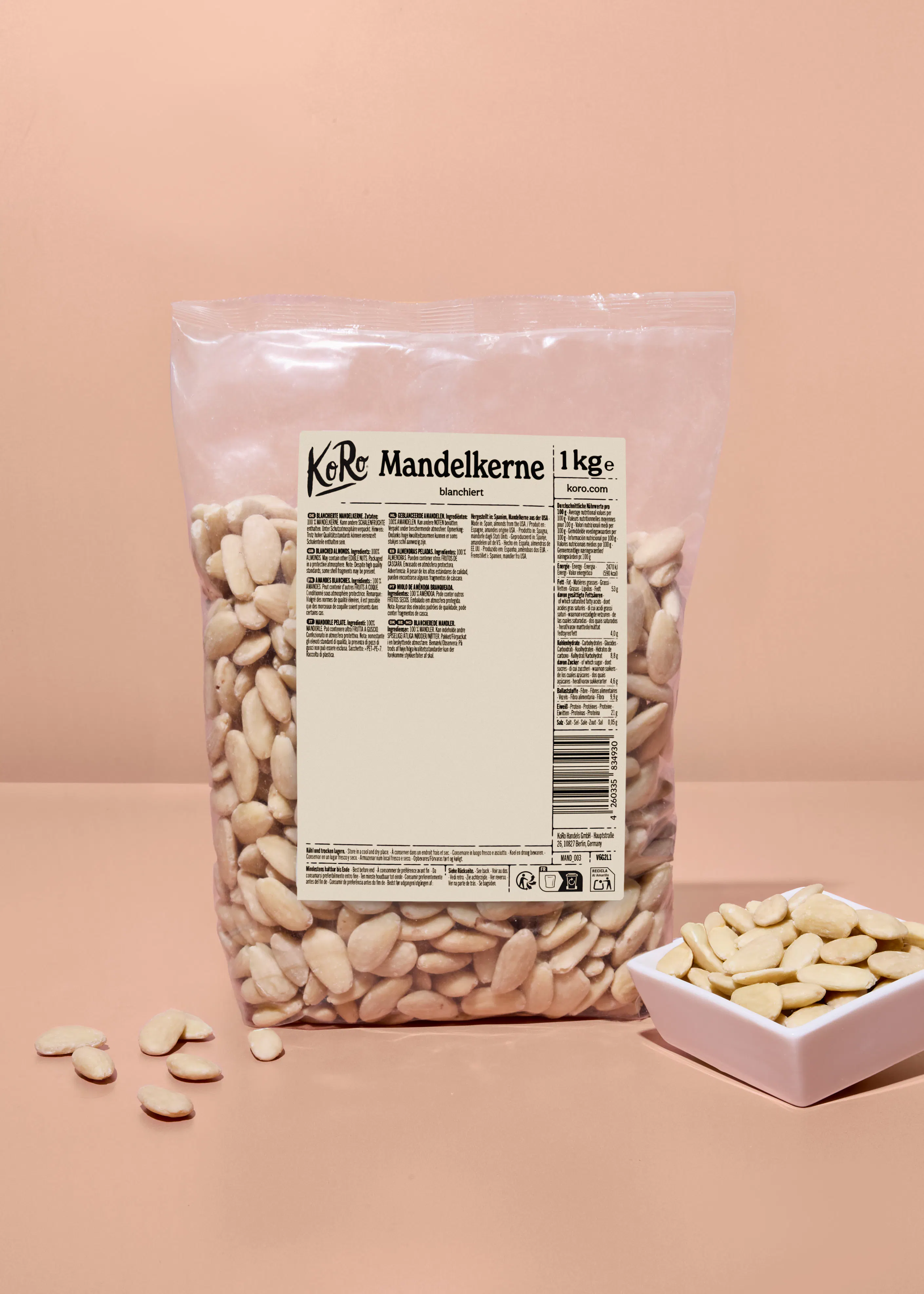 eine durchsichtige plastiktüte mit 1 kg blanchierten mandelkernen der marke koro steht auf einer rosa oberfläche, daneben eine kleine weiße schale mit weiteren mandelkernen und einige lose mandeln.