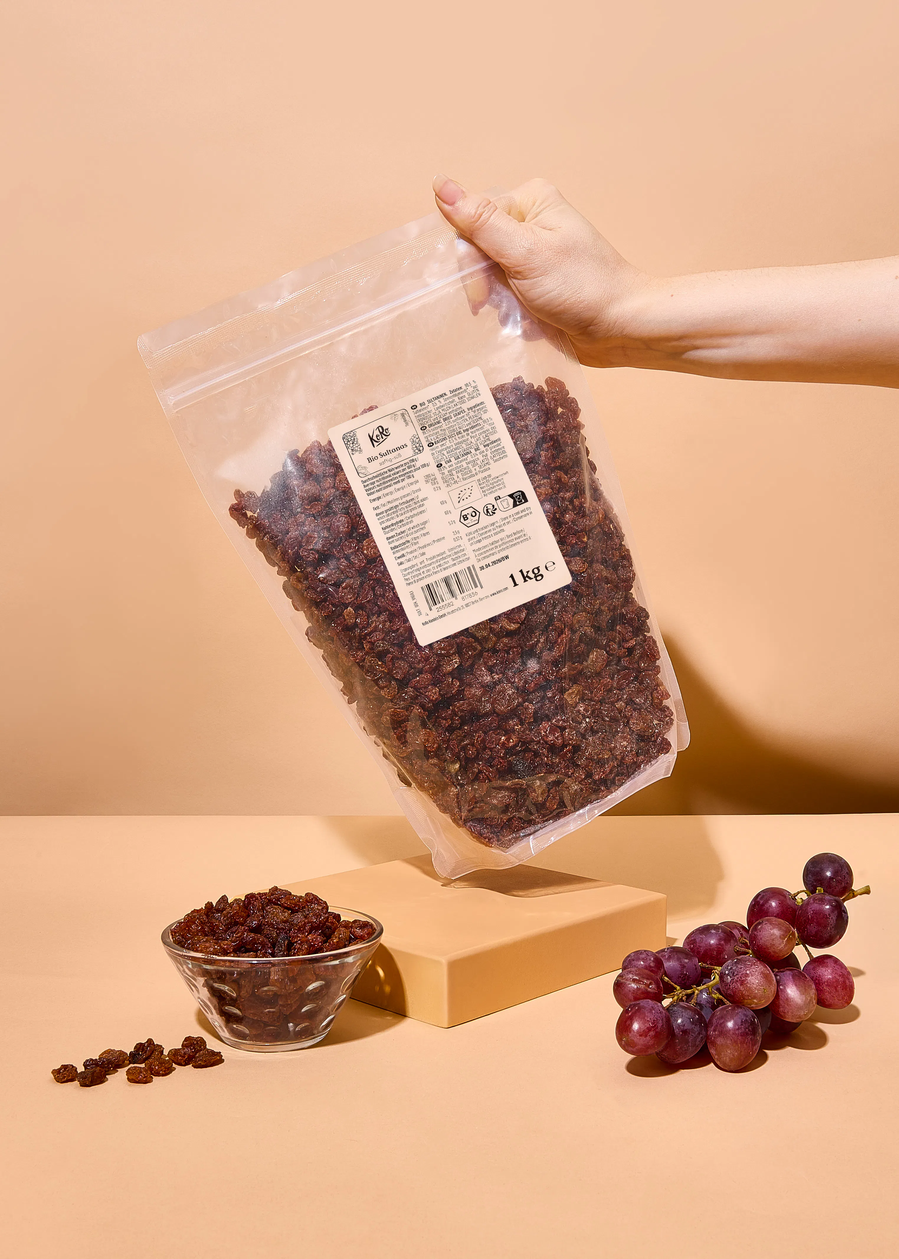 eine hand hält einen transparenten beutel mit 1 kg bio-sultaninen, daneben stehen eine schale mit sultaninen und eine traube auf einem beigen untergrund.