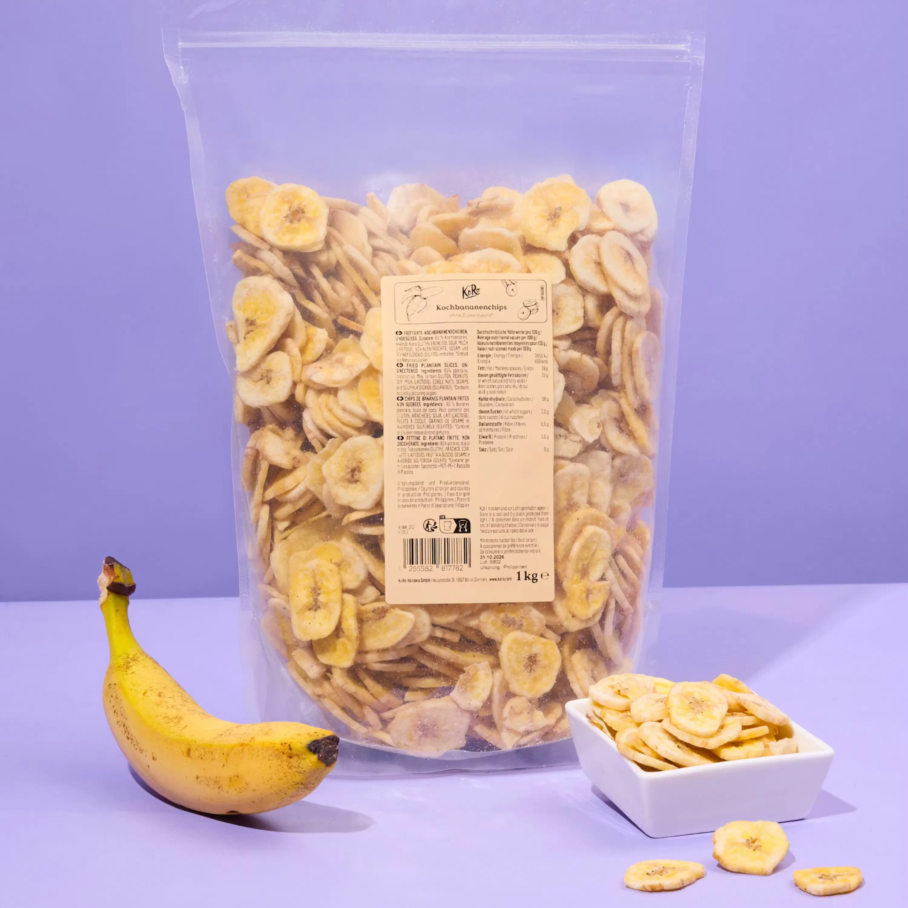 un grand sac transparent de chips de banane, une banane fraîche et un petit bol blanc rempli de chips de banane, le tout sur un fond violet.