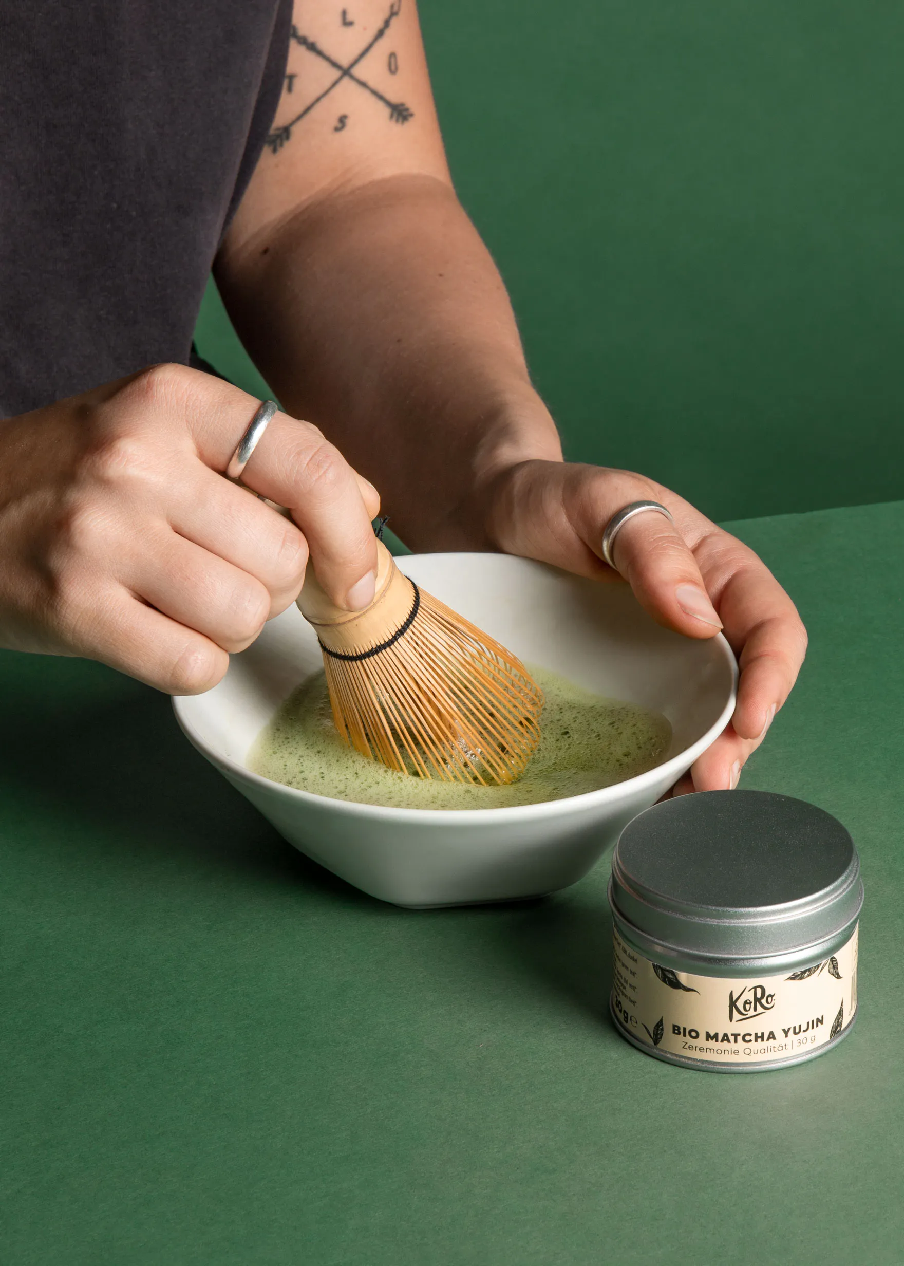 eine Person rüscht eine weiße Schüssel mit grünem Tee neben einer Dose bio matcha yujin