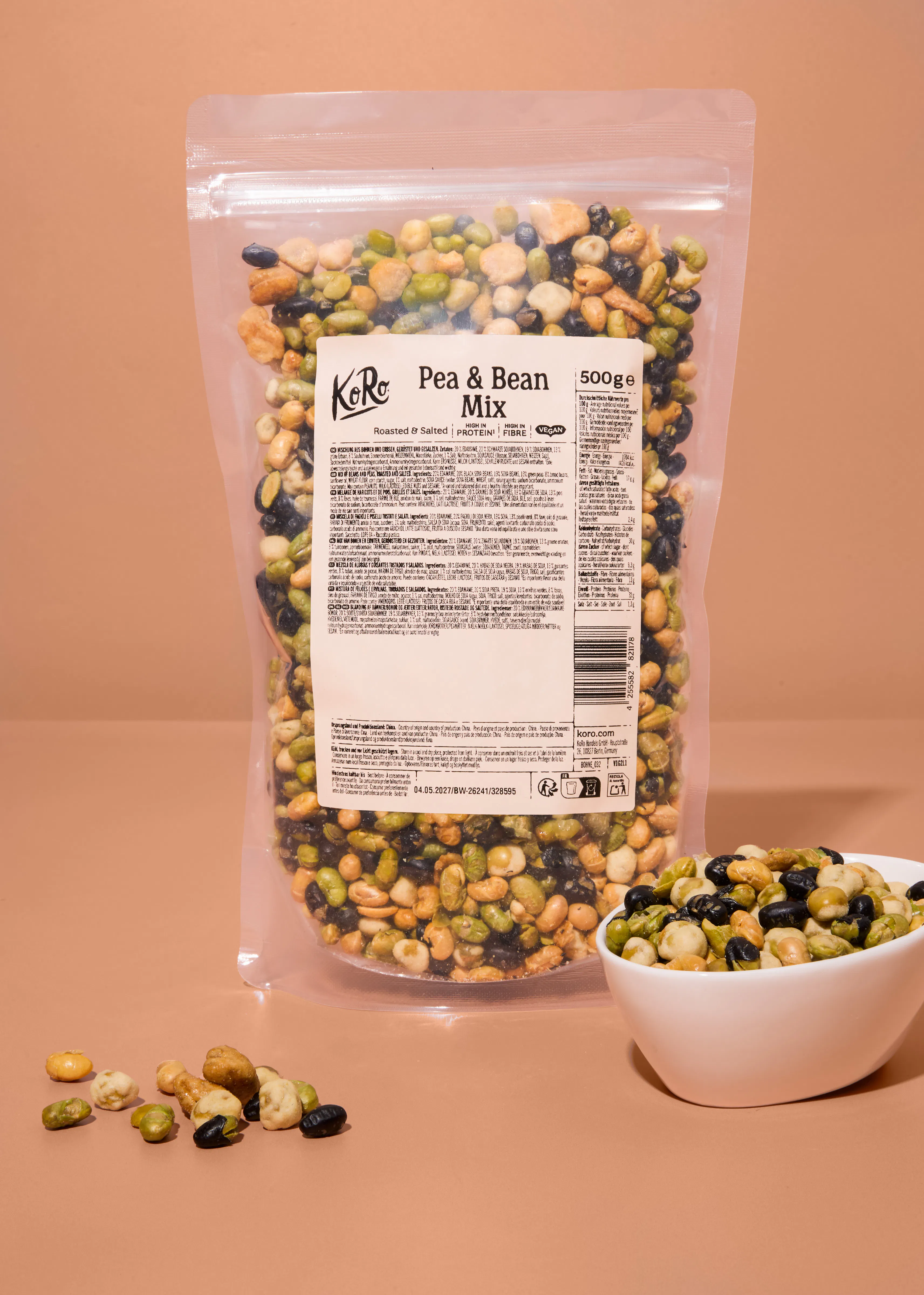 eine durchsichtige verpackung mit koro pea & bean mix, bestehend aus gerösteten und gesalzenen erbsen und bohnen, steht neben einer weißen schale mit einer kleinen portion des snacks vor einem beigen hintergrund.
