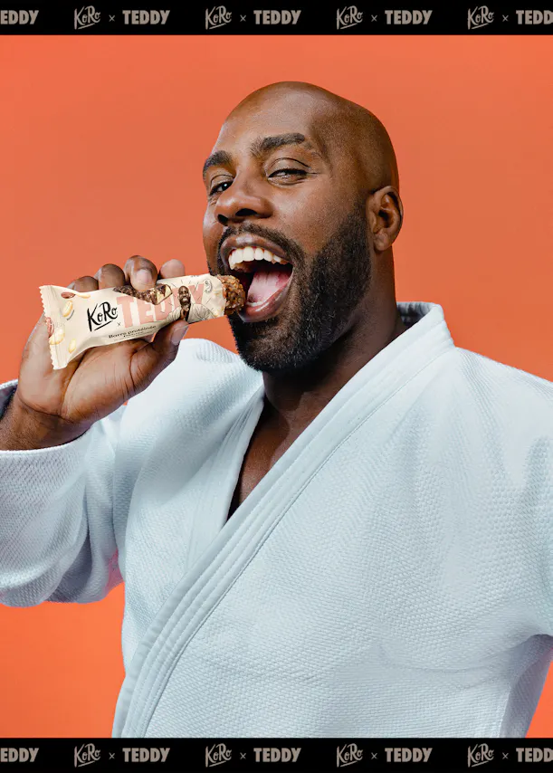 un homme barbu en kimono blanc mangeant une barre protéinée koro x teddy, la bouche ouverte et l'œil droit cligné, sur un fond orange.