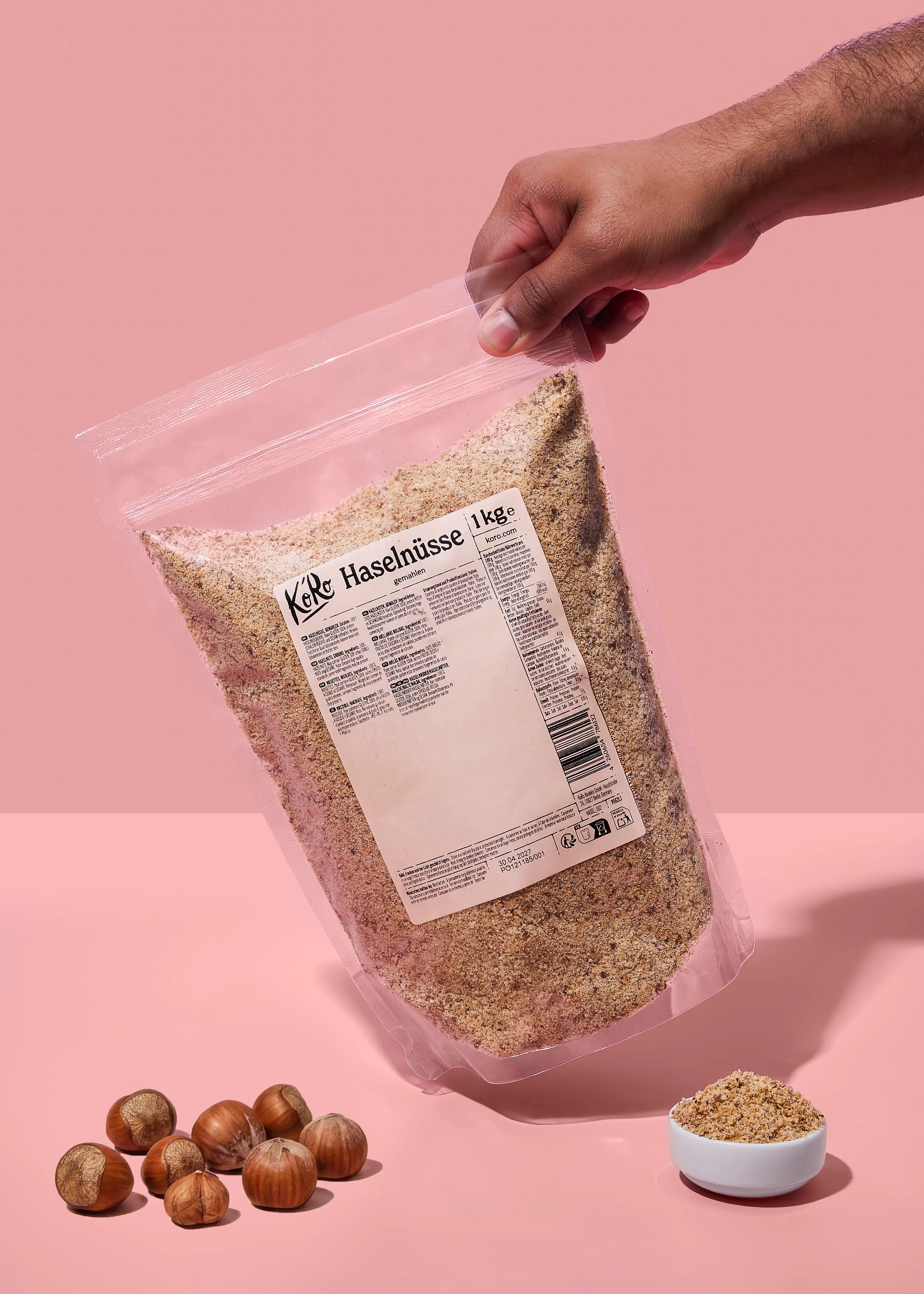una mano tiene una busta trasparente di nocciole macinate da 1 kg, con nocciole intere e una ciotola di nocciole macinate su uno sfondo rosa.