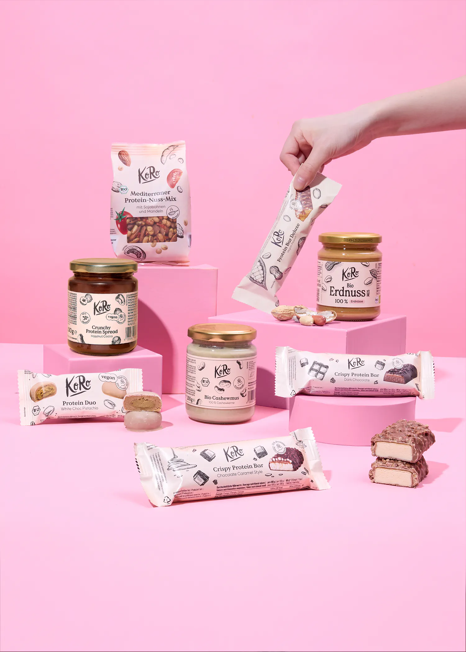 une collection de produits protéinés koro, comprenant des pâtes à tartiner, des mélanges de noix et des barres, disposés sur des socles roses sur un fond rose, avec une main tenant une barre protéinée.