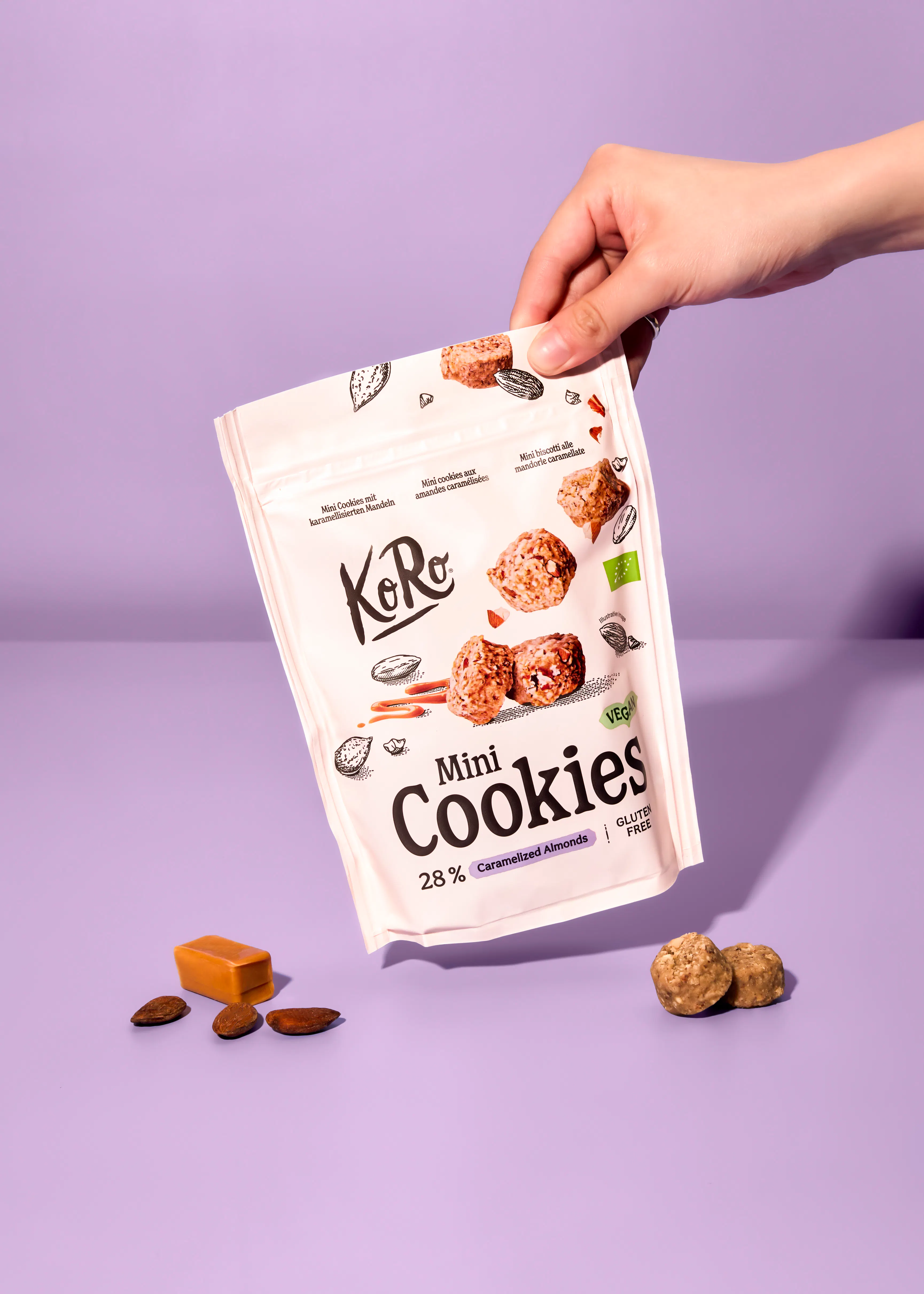 eine hand hält eine packung koro mini cookies mit karamellisierten mandeln, die vegan und glutenfrei sind, während einige kekse, mandeln und karamellstücke auf einem lila hintergrund verstreut sind.