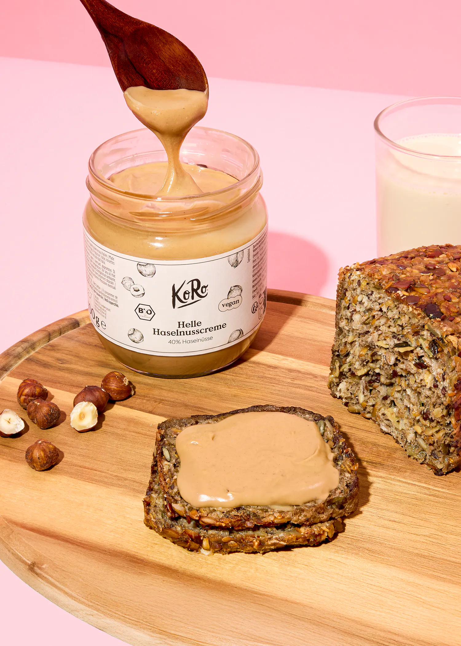 ein holzlöffel tropft helle haselnusscreme aus einem glas auf ein holzbrett, das auch eine bestrichene brotscheibe, einen brotlaib, ganze haselnüsse und ein glas milch zeigt.