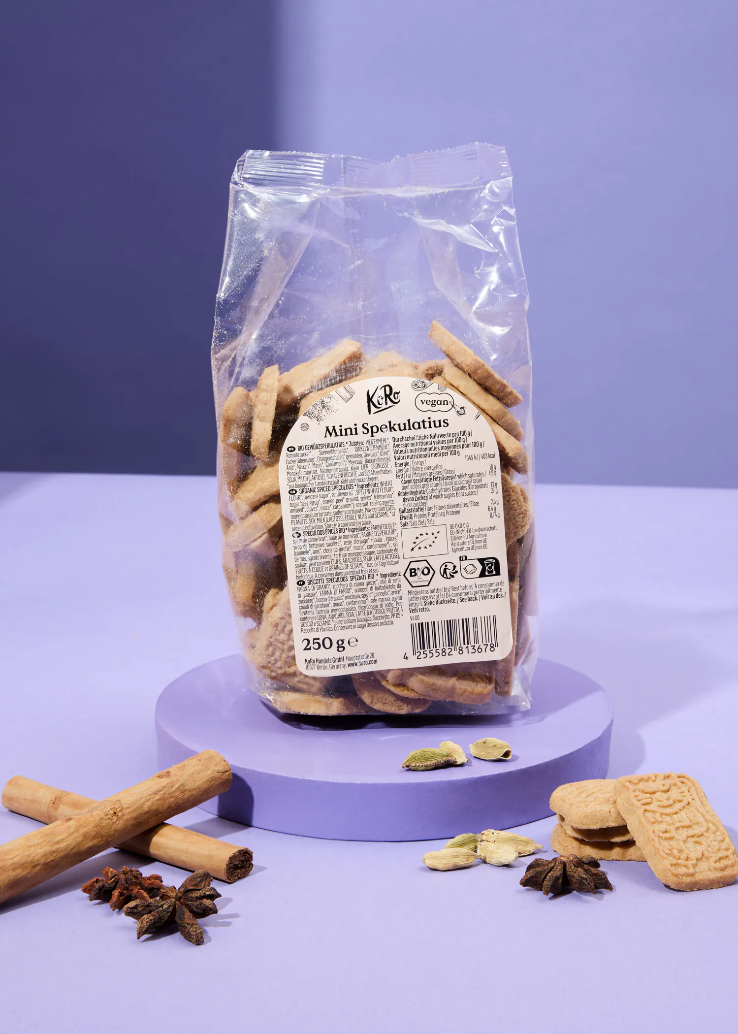 a clear plastic bag of vegan mini spekulatius cookies rests on a purple stand, surrounded by cinnamon sticks, star anise, cardamom pods, and a few loose cookies.
