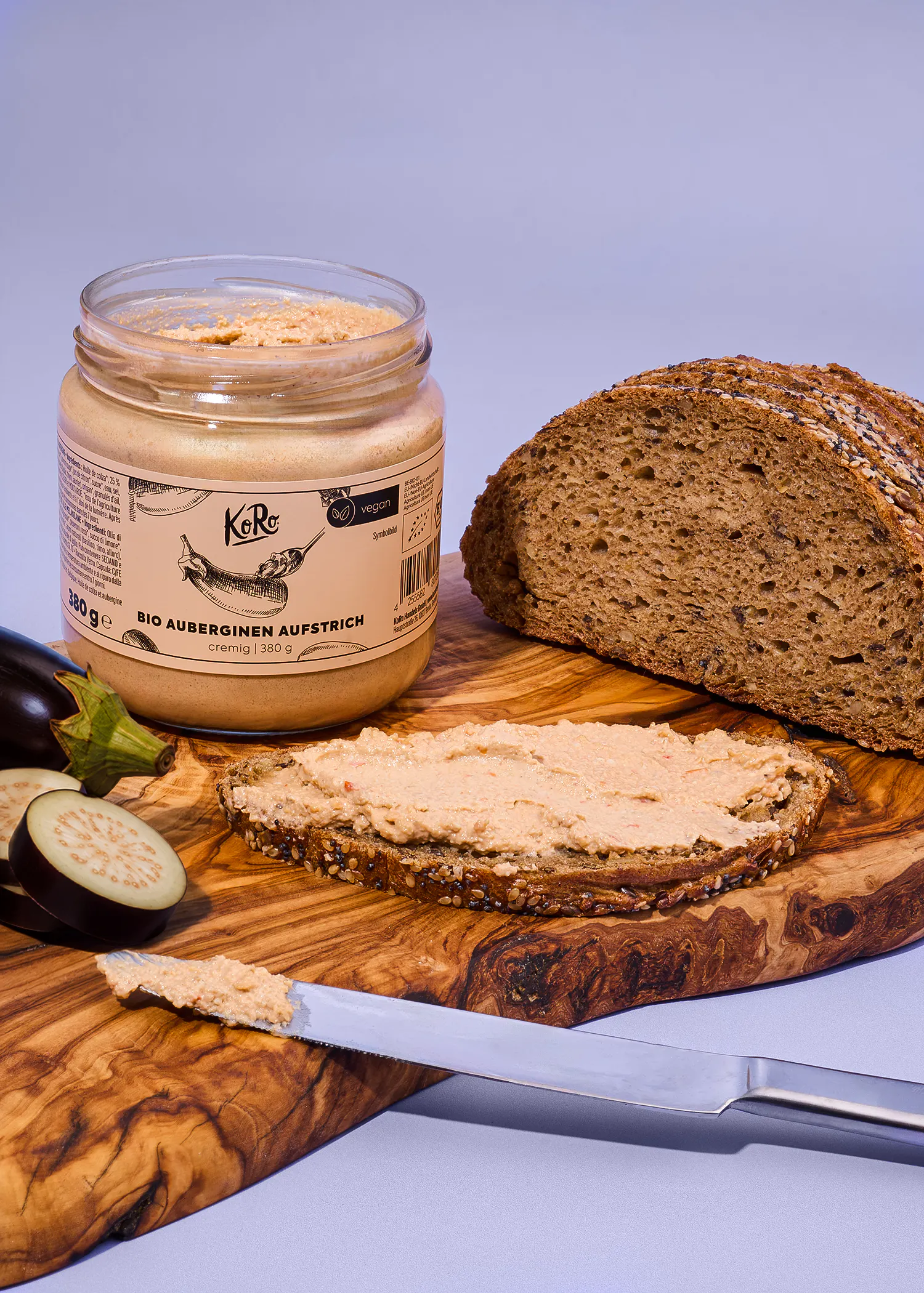ein glas koro bio-auberginen-aufstrich steht neben einer scheibe brot mit aufstrich und einem messer auf einem holzbrett.