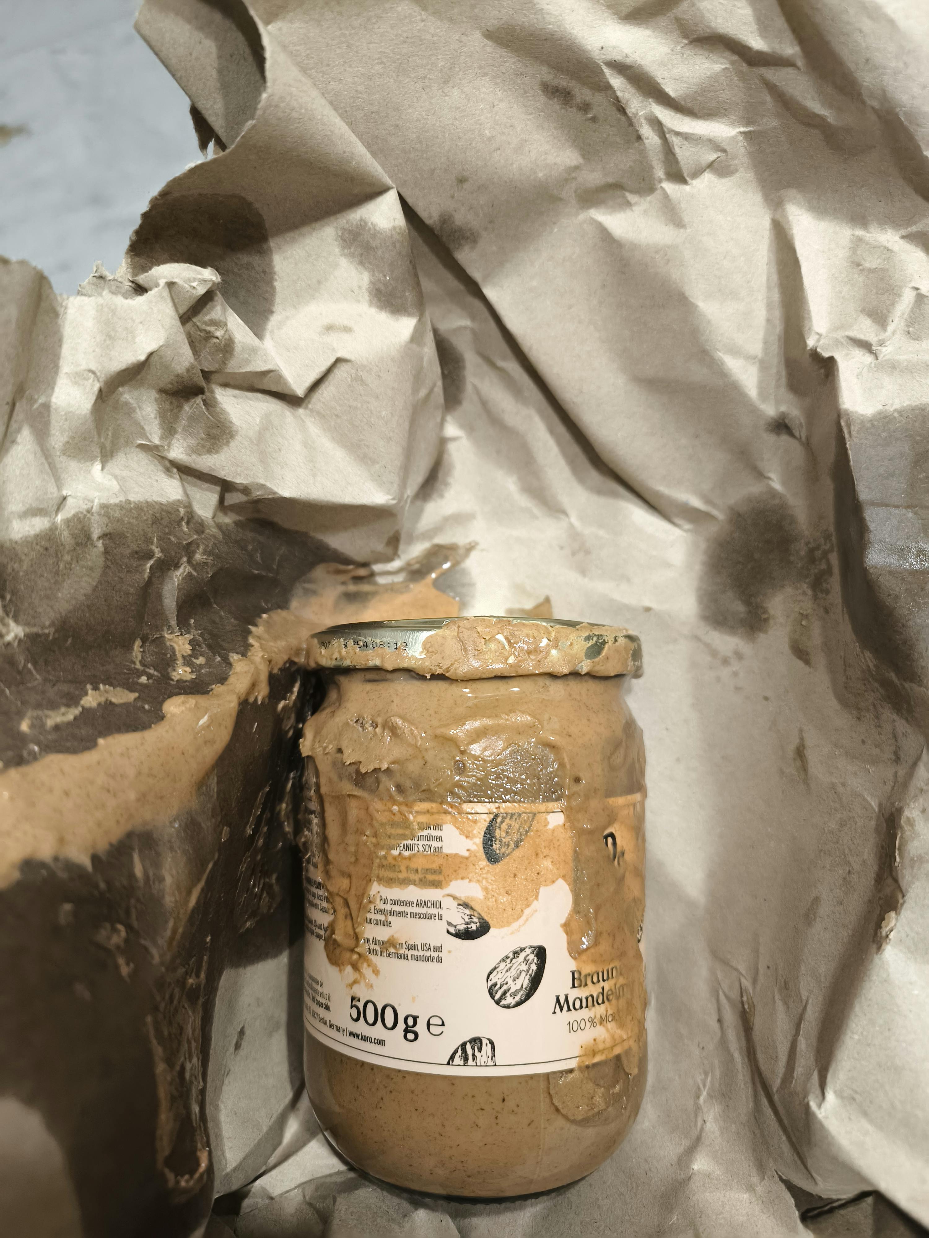 ein glas braunes mandelmus, das außen mit mandelmus verschmiert ist und in zerknittertem, braunem papier liegt.