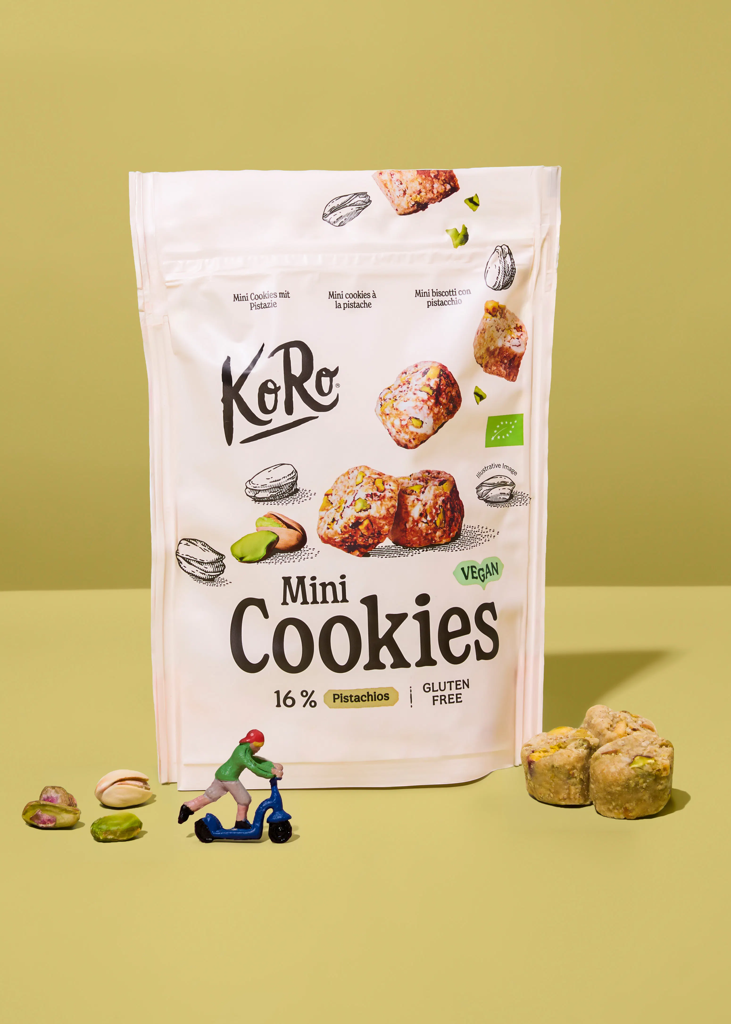 eine weiße verpackung von koro mini cookies mit pistazien, daneben liegen lose pistazien, kekse und eine kleine figur auf einem roller auf hellgrünem hintergrund.