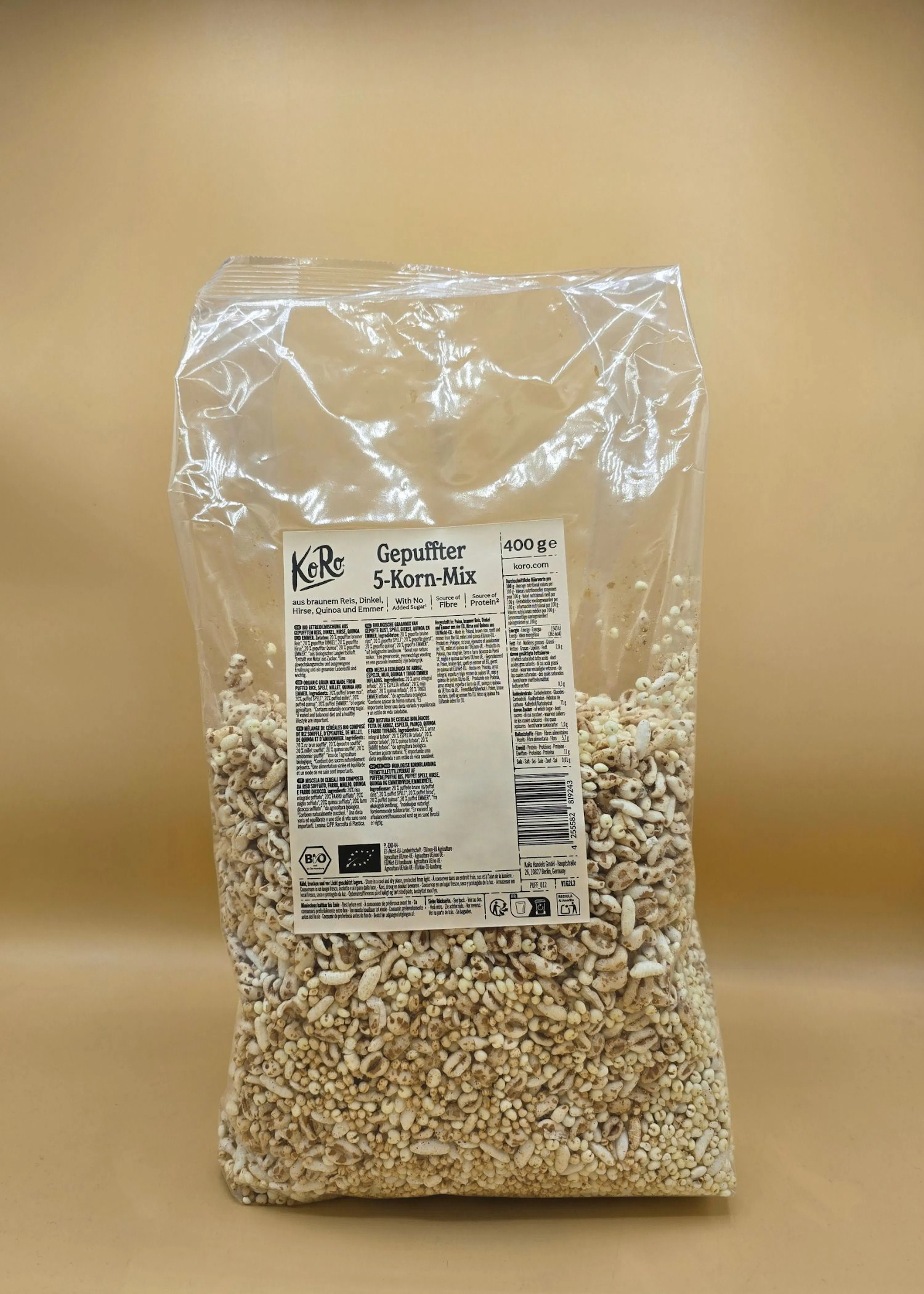 a clear plastic bag of koro gepuffter 5-korn-mix (puffed 5-grain mix) stands upright, filled with light brown and white puffed grains, against a plain background.