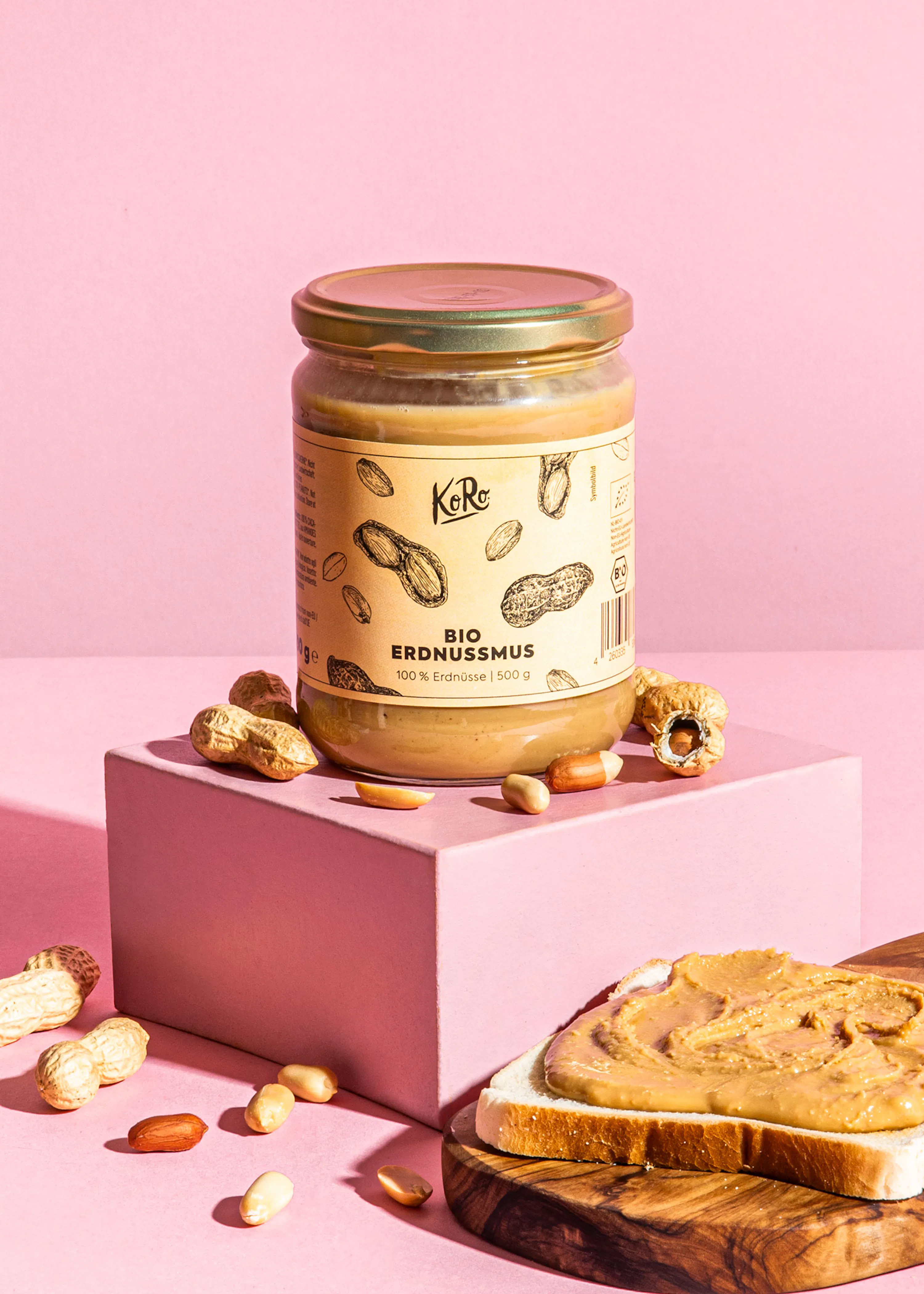 a jar of ko ro bio erdnussmus peanut butter sits on a pink block, accompanied by loose peanuts and a slice of toast spread with peanut butter.