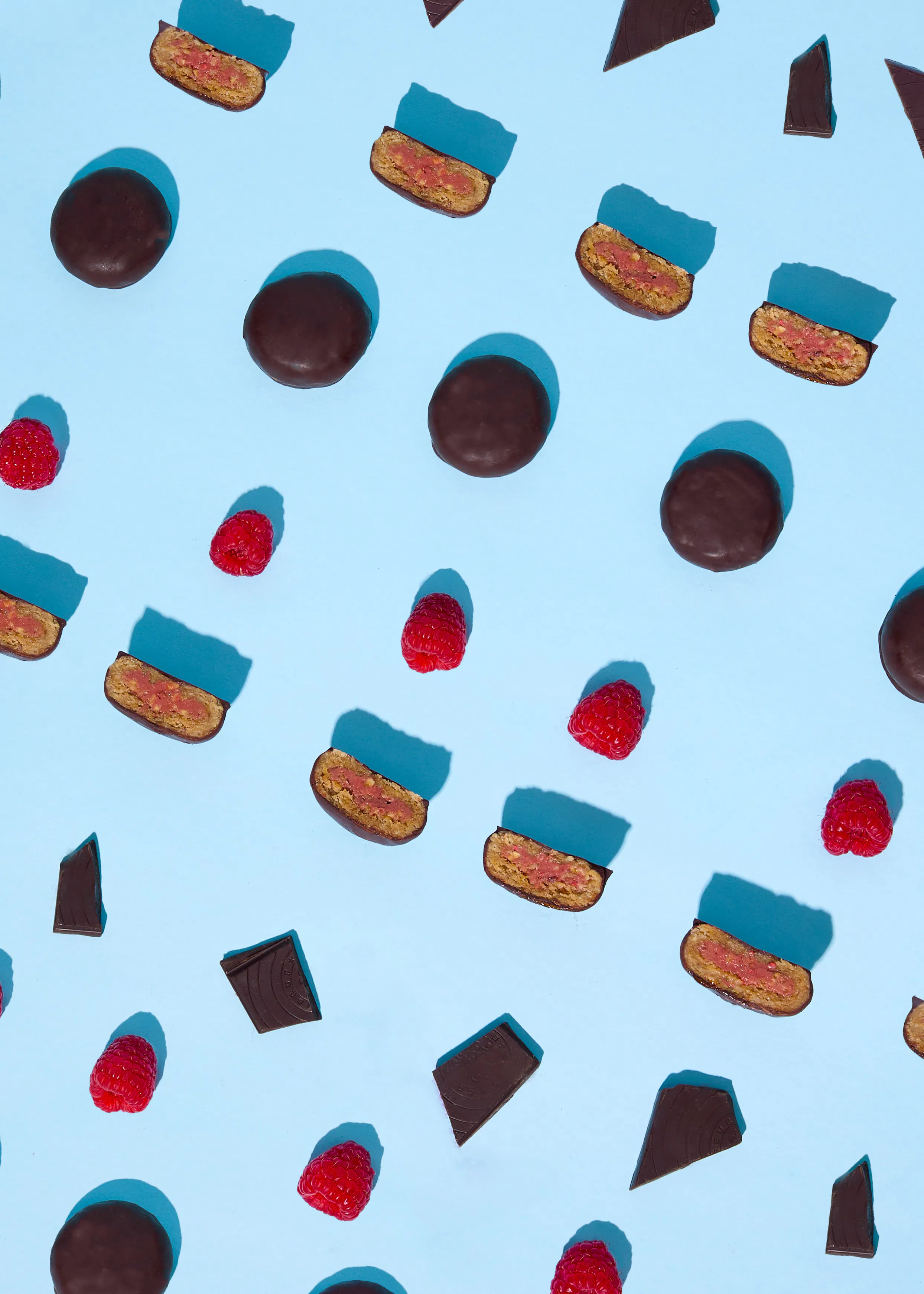 ein muster aus schokoladenkeksen, einigen halbiert mit rosa füllung, frischen himbeeren und schokoladenstücken auf hellblauem hintergrund mit scharfen schatten.