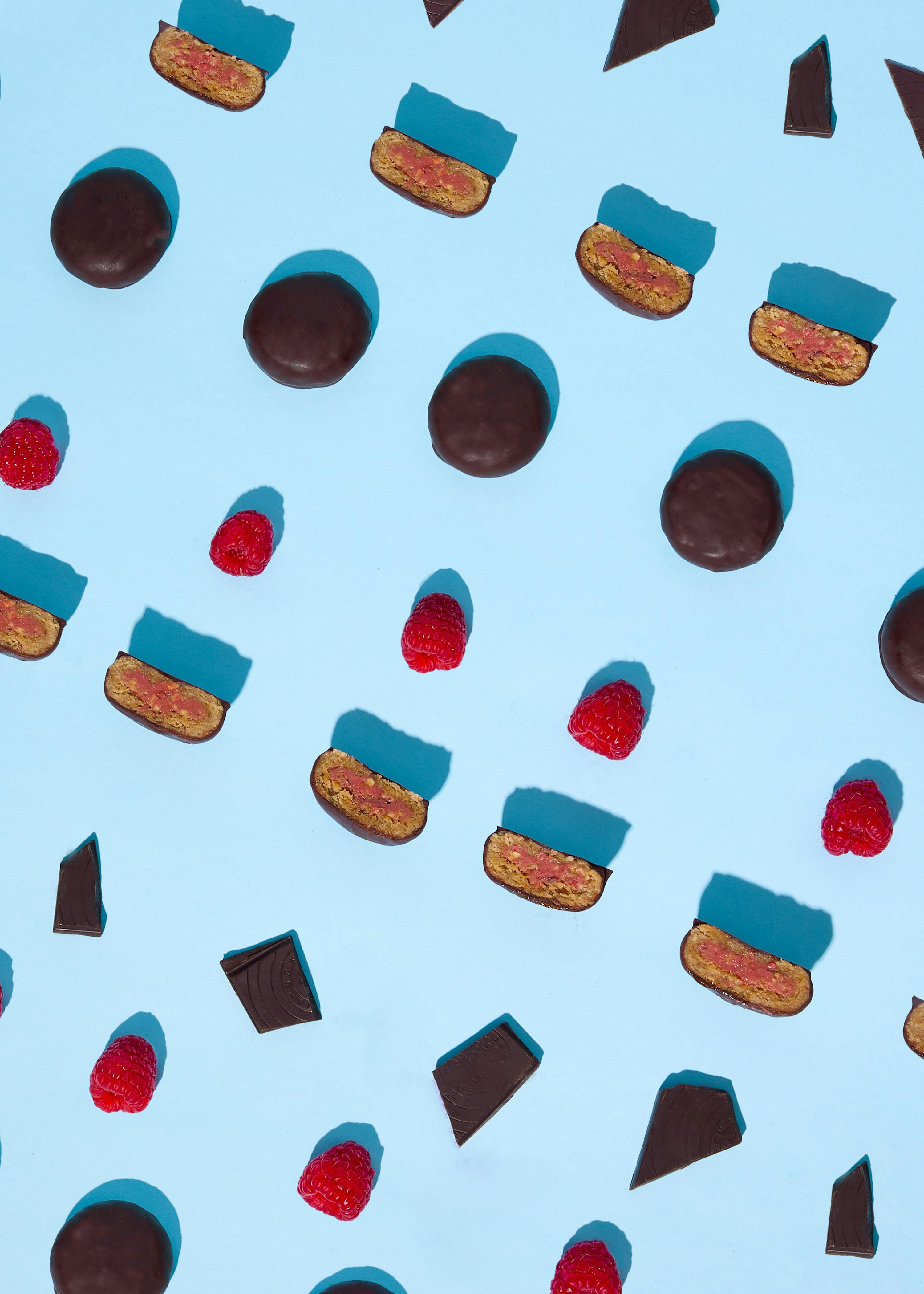 un motivo di biscotti ricoperti di cioccolato, alcuni interi e altri tagliati a metà con ripieno rosa, lamponi freschi e pezzi di cioccolato fondente sparsi su uno sfondo azzurro con ombre nette.