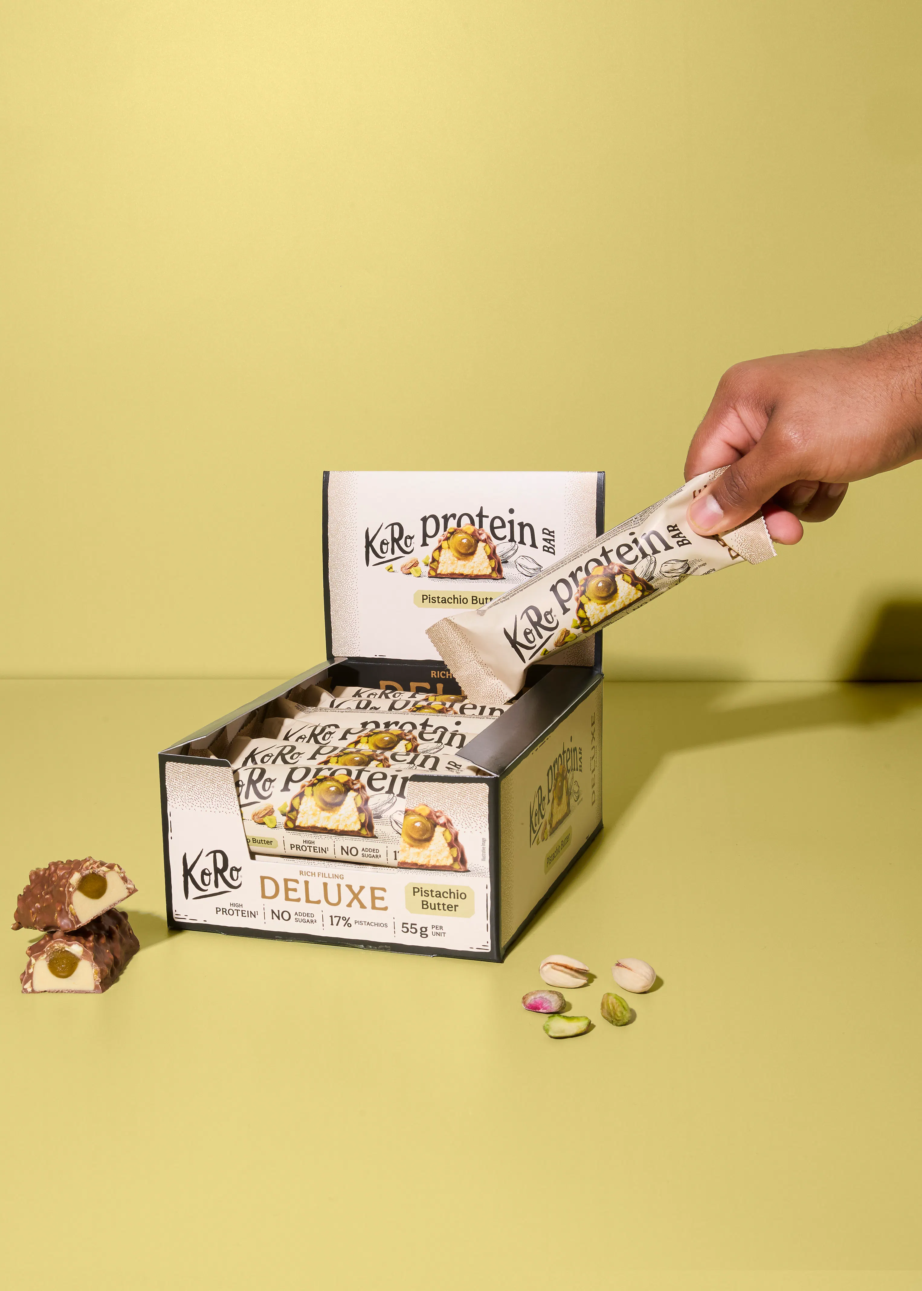 una mano prende una barretta proteica al burro di pistacchio koro deluxe da una scatola, con una barretta tagliata e pistacchi sparsi in primo piano.