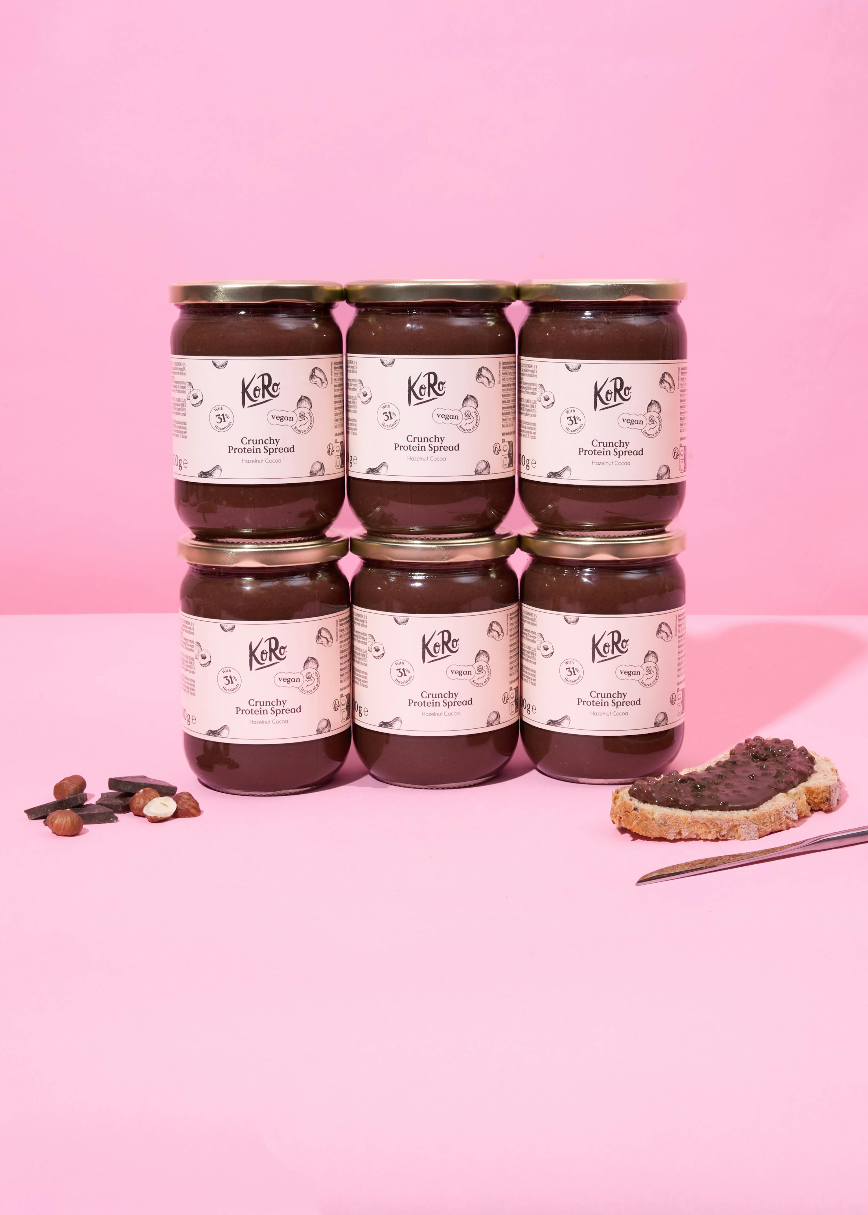 six jars of koro crunchy protein spread hazelnut cocoa are stacked on a pink surface next to loose hazelnuts, chocolate pieces, and a slice of bread with the spread.