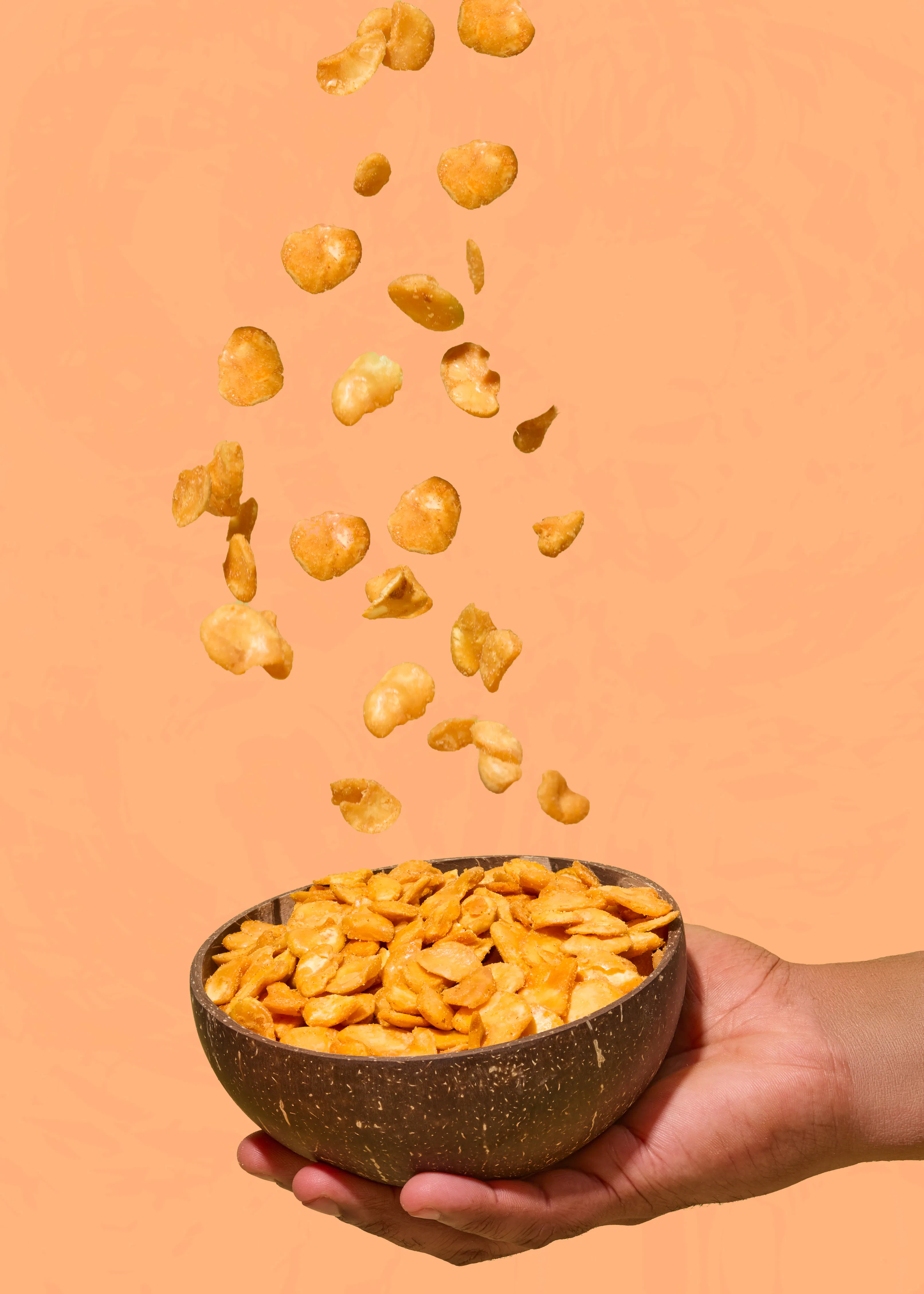 une main tient un bol en noix de coco rempli de fèves frites croustillantes avec d'autres fèves qui tombent en suspension sur un fond orange.