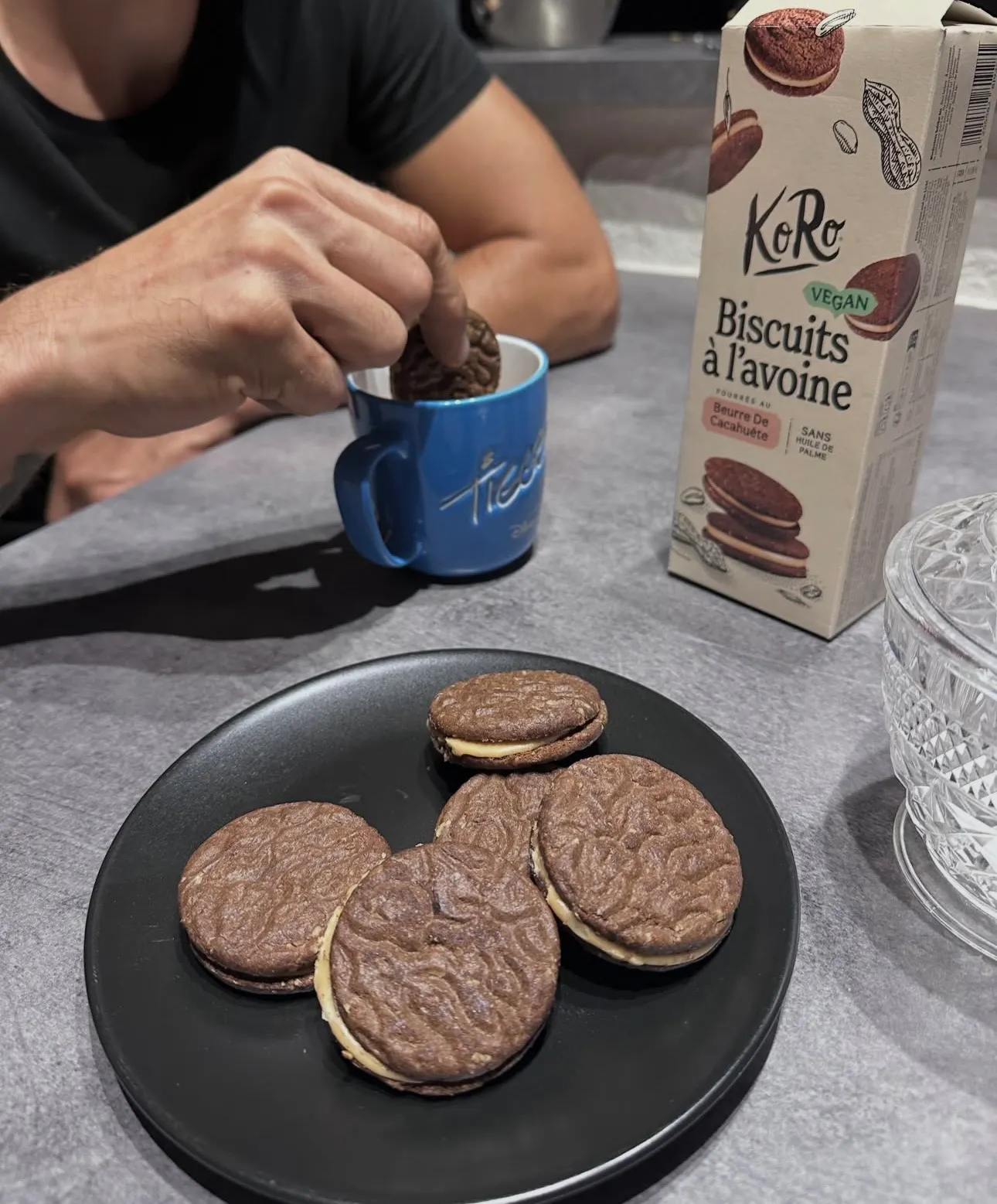 eine person tunkt einen koro-haferkeks mit erdnussbutterfüllung in eine blaue tasse, während weitere kekse auf einem schwarzen teller liegen.